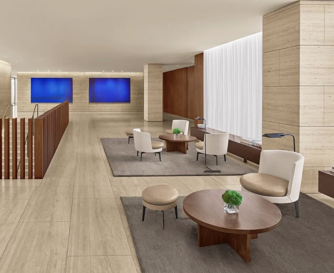 Modern hotel lobby with seating area, white chairs, round wooden tables with green plants, beige tiled floors, large white curtain, two blue screens on wall.