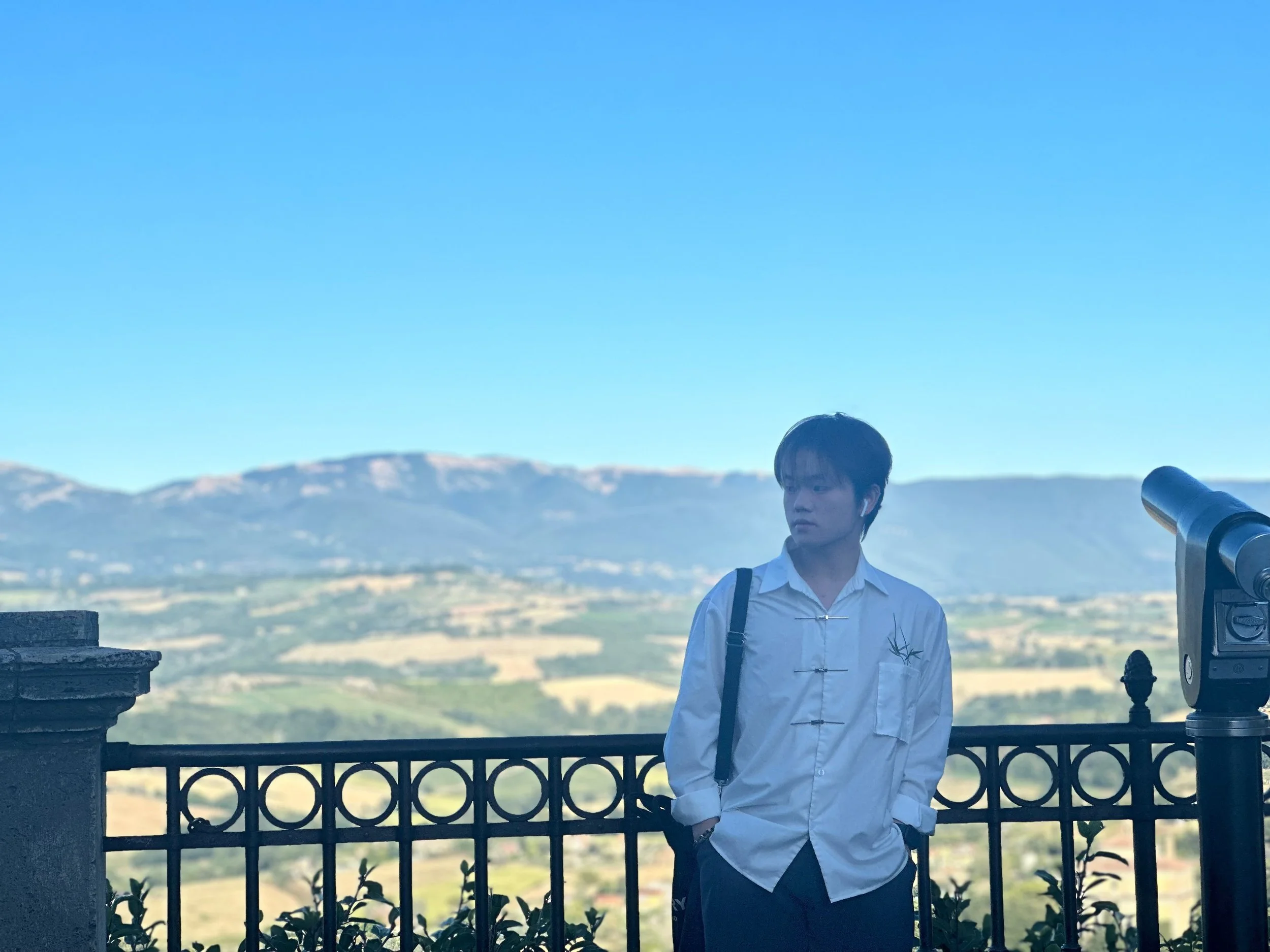 A young man with black hair, wearing a white shirt and black pants, stands near a telescope on a balcony with a scenic mountain landscape in the background.