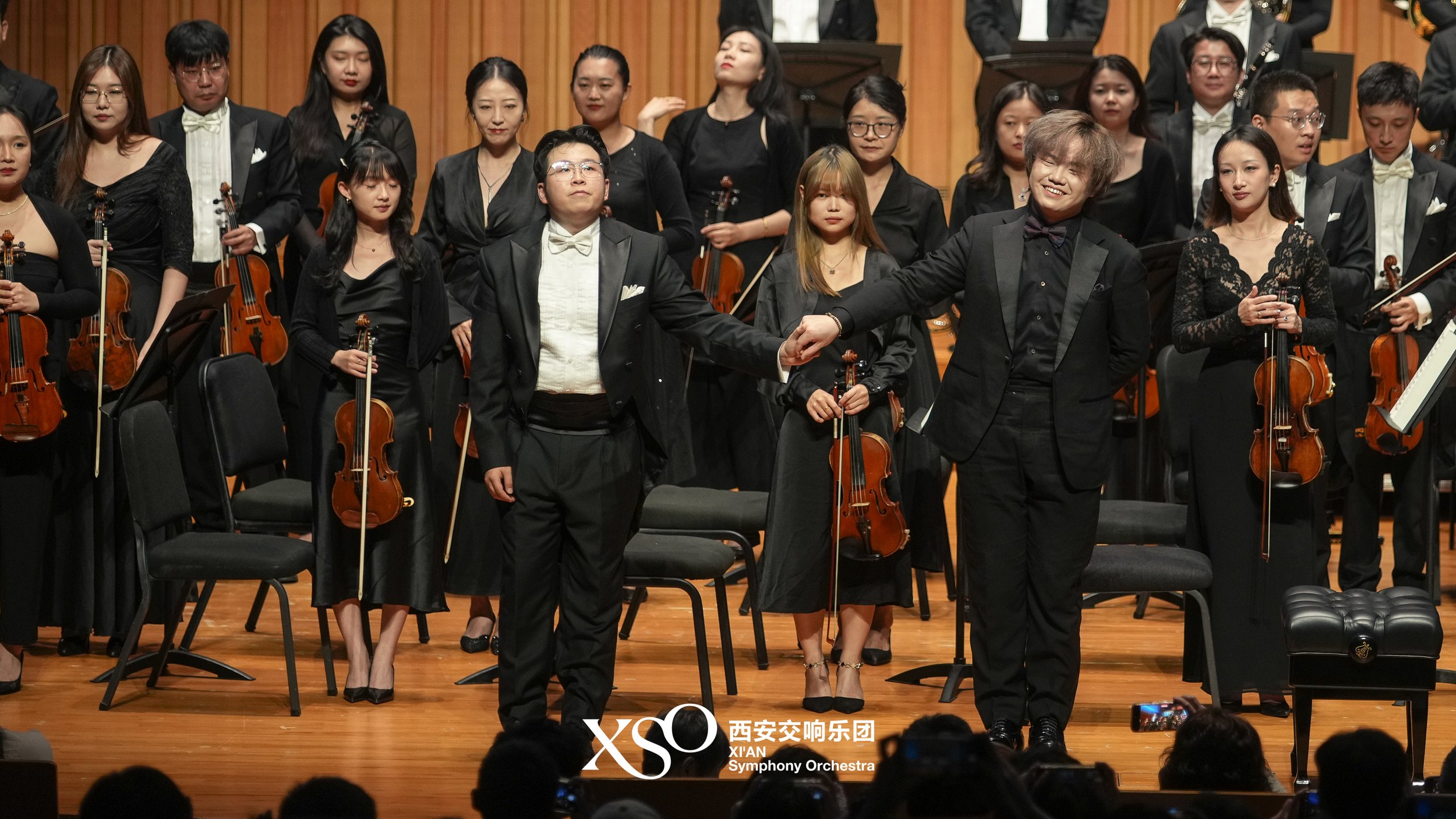 Orchestra members in formal black attire on stage, holding violins, with two conductors in front holding hands, during a symphony performance.