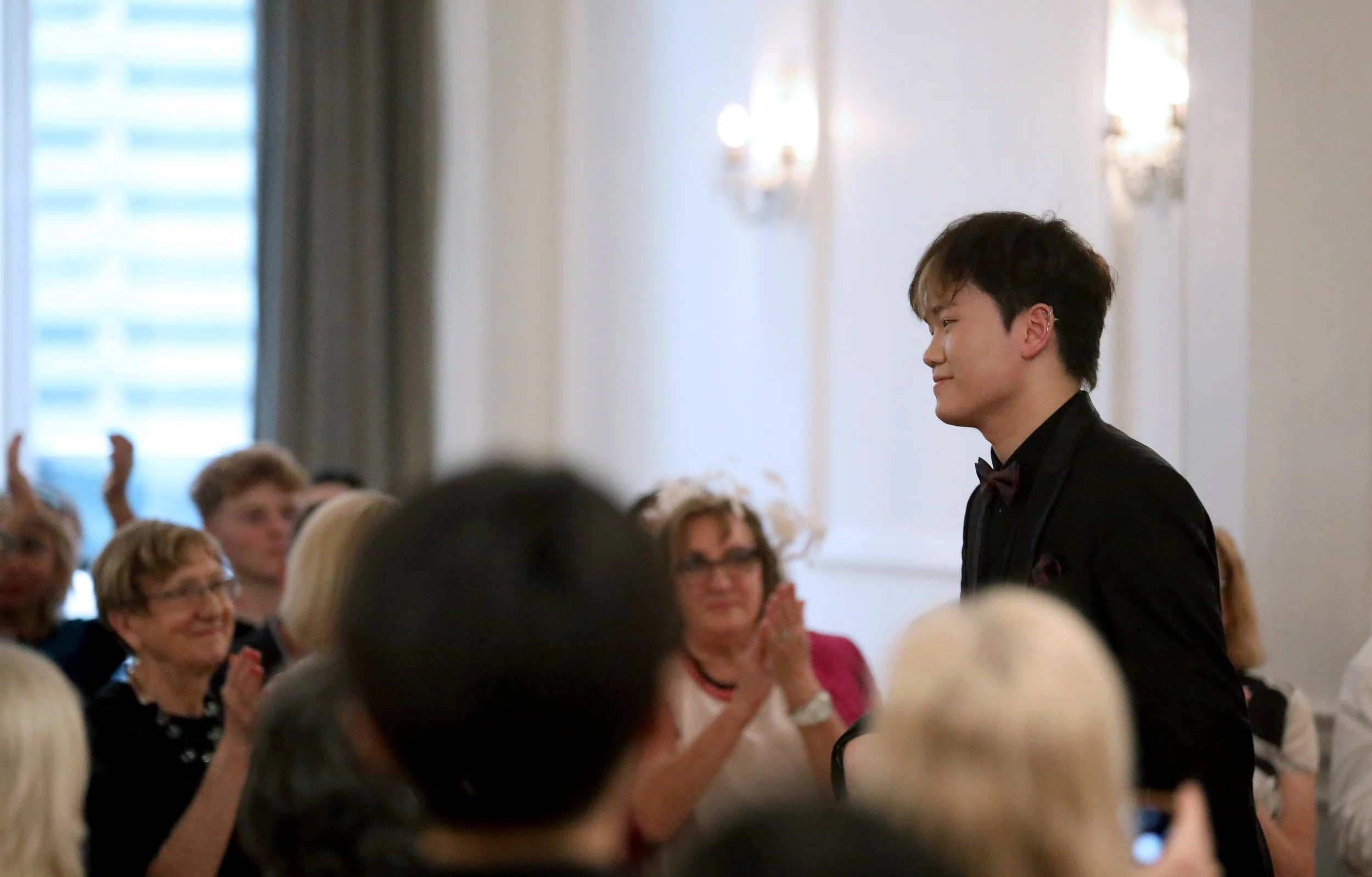 Hao Rao performing recital in London. A young man in a black suit and bow tie stands smiling in a formal event, with an audience clapping and smiling in a bright, elegant room.