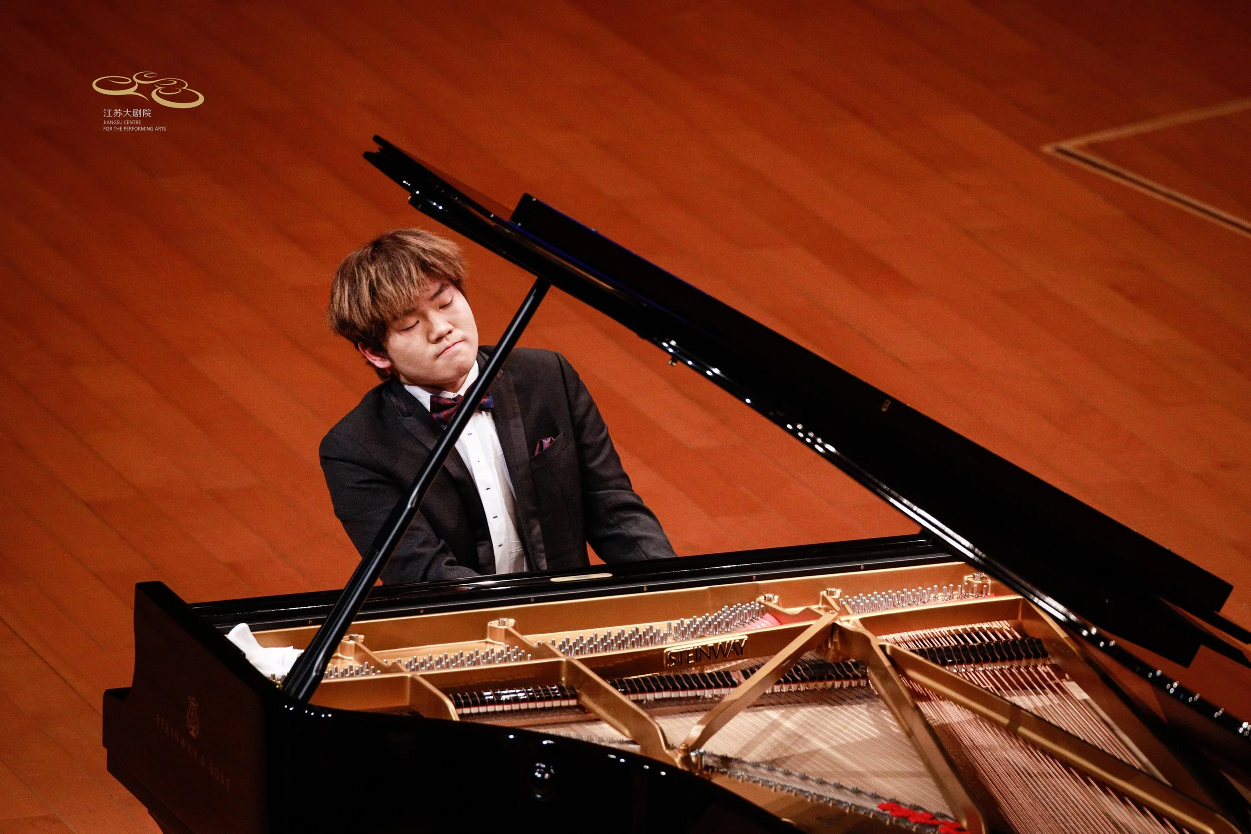 Hao Rao A young man playing a grand piano on a wooden stage, wearing a tuxedo with a bowtie, with his eyes closed and an expression of concentration.