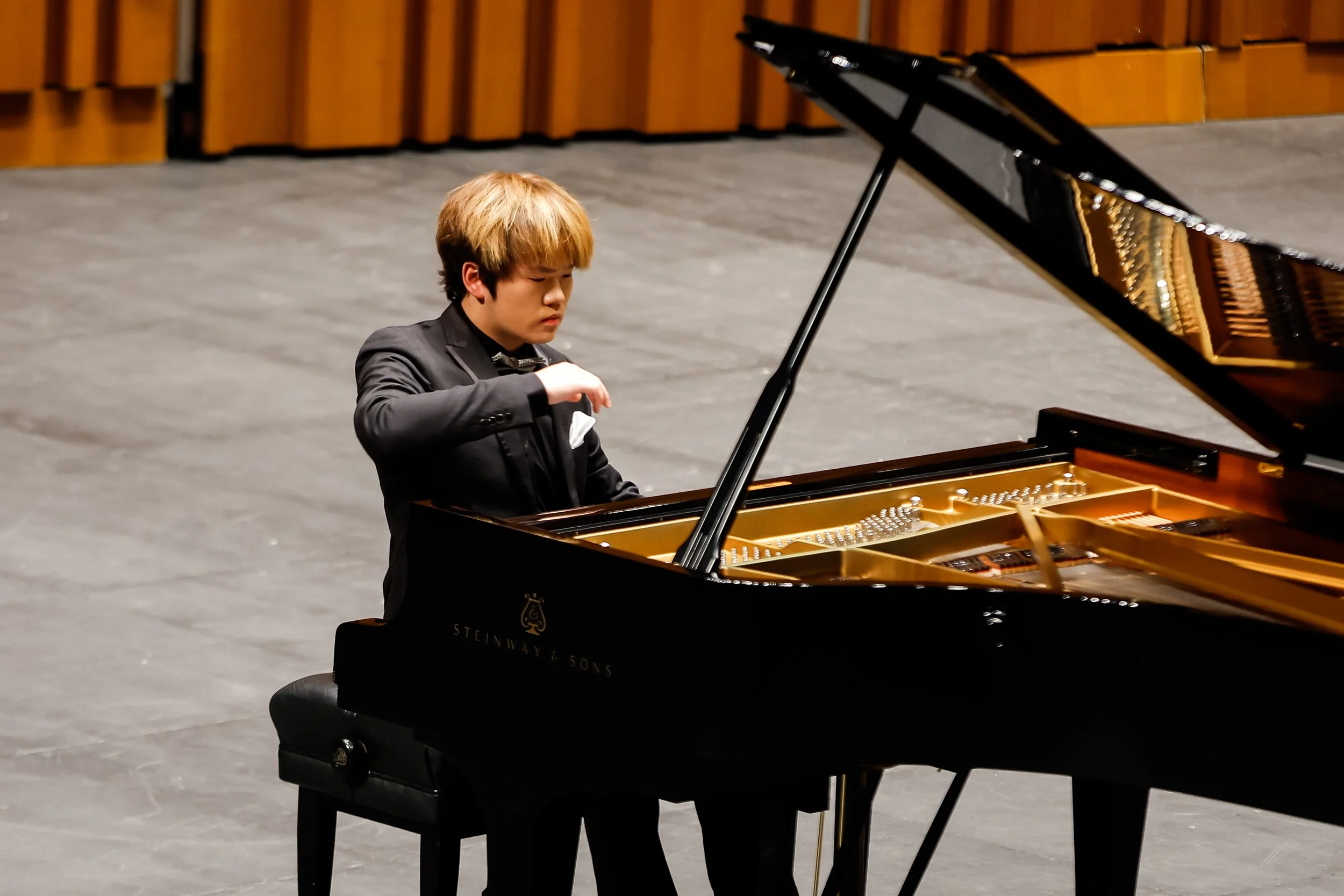 A young man in a black suit playing a grand piano on stage.