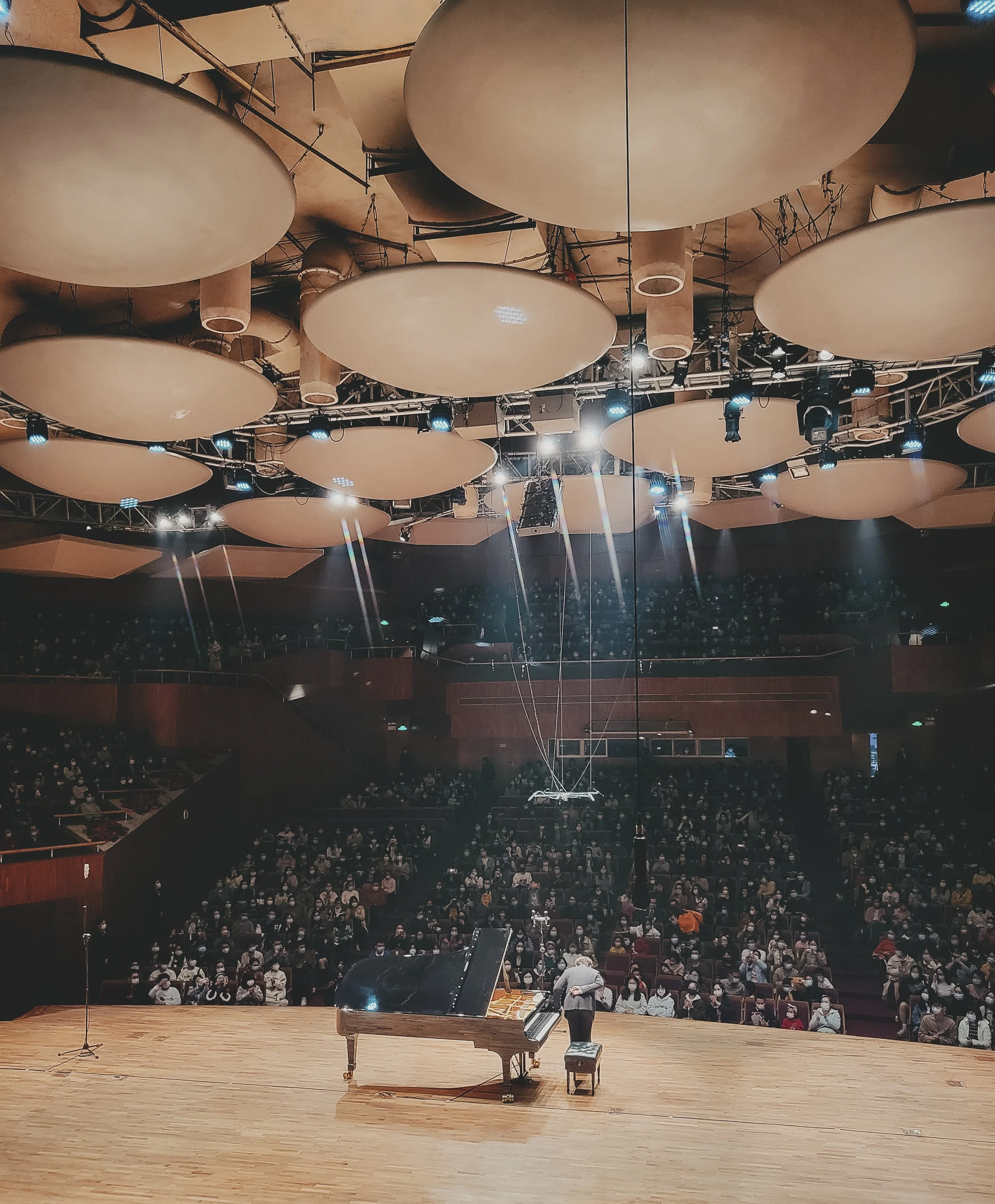 View of an empty stage in a concert hall with a grand piano, a stool, and a microphone, facing an audience with multiple tiers of seating, large circular acoustic panels hanging from the ceiling, and stage lights overhead.