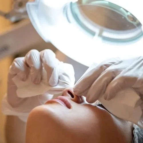 Procedimento de atendimento dermatológico ou estético, com profissional usando luvas e lupa, focado na área do rosto.