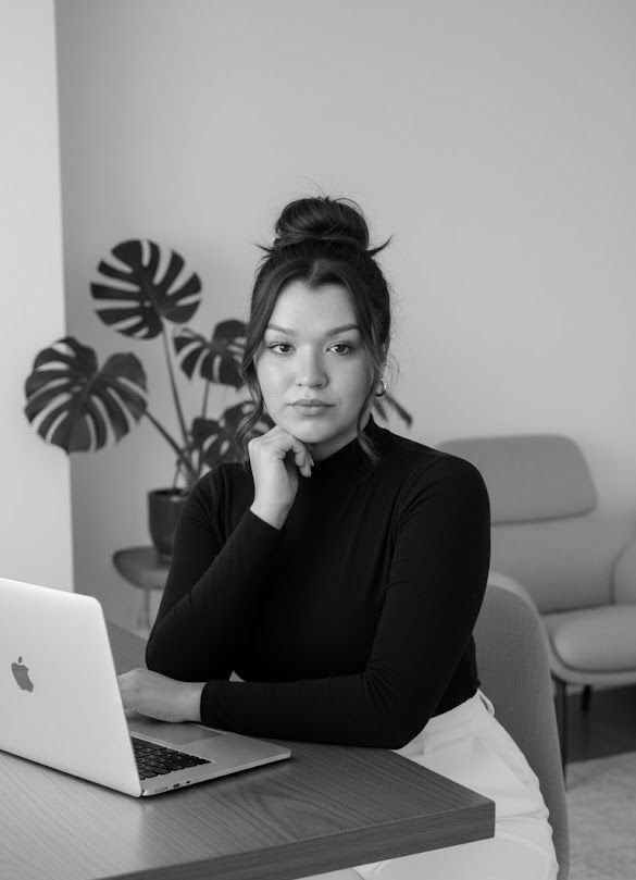 Mulher sentada à mesa com um laptop Apple, cabelo em coque, expressão pensativa, fundo com plantas decorativas e sofá