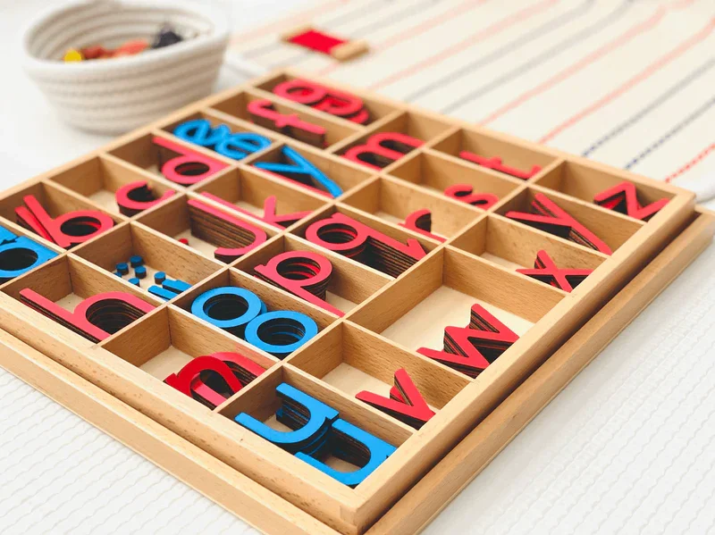 Wooden Scrabble-like letter tiles in a grid with some letters in red and blue, and a bowl of small black and yellow tokens in the background.