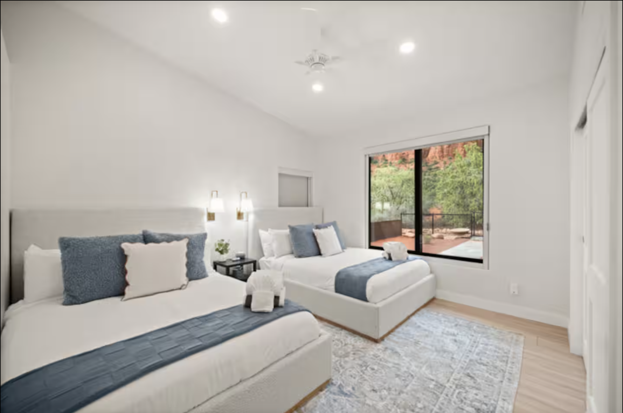 Two white beds with blue pillows and blankets, nightstand between them, view of a sliding glass door leading to an outdoor balcony with red rock and green trees outside.