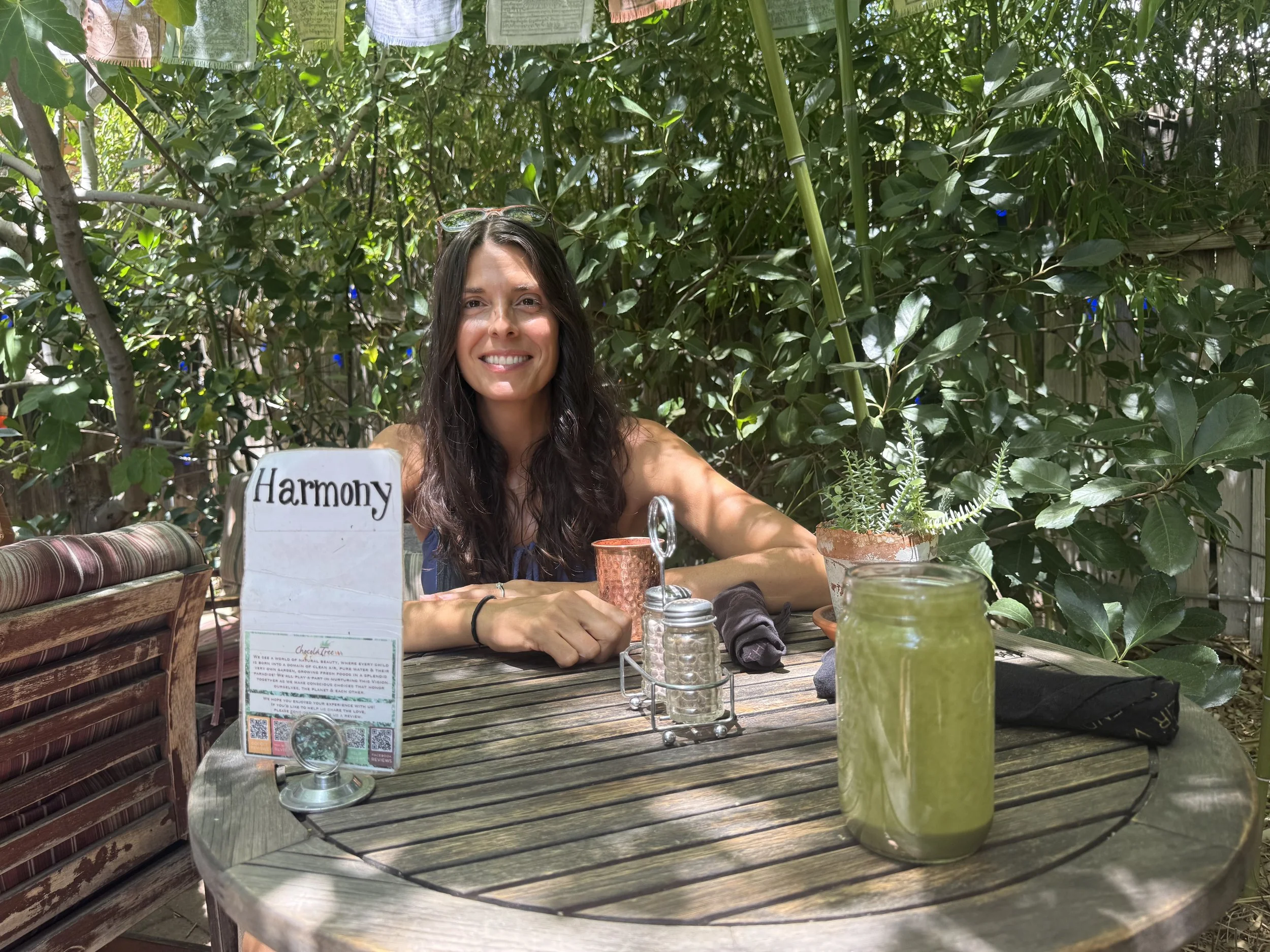 Smiling woman with long dark hair sitting at a wooden outdoor table surrounded by green foliage, with drinks and a plant on the table, and a small sign that says 'Harmony.'
