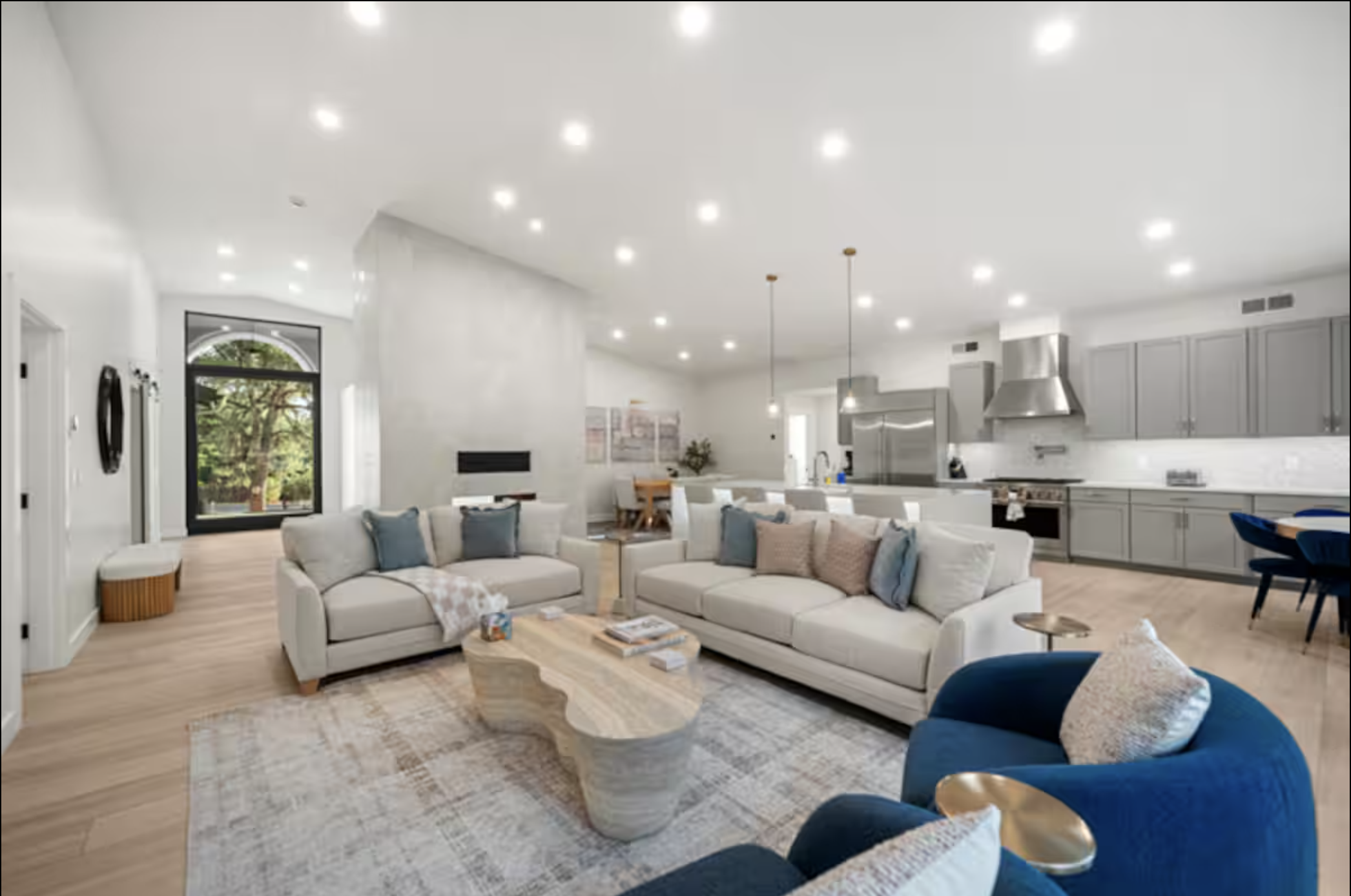 Open-concept living room with white sofas, a blue armchair, a stone coffee table, and a kitchen with gray cabinets and stainless steel appliances.