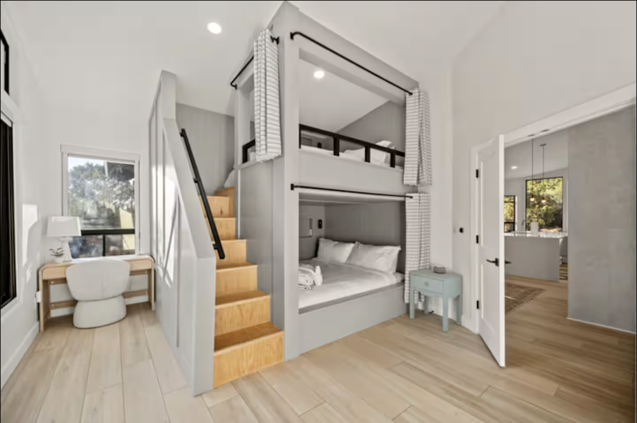 Interior of a modern, light-colored bedroom with a small desk and chair near a window, a loft bed with a staircase, and a built-in bed beneath the loft with pillows and bedding. The overall style is airy and minimalist.