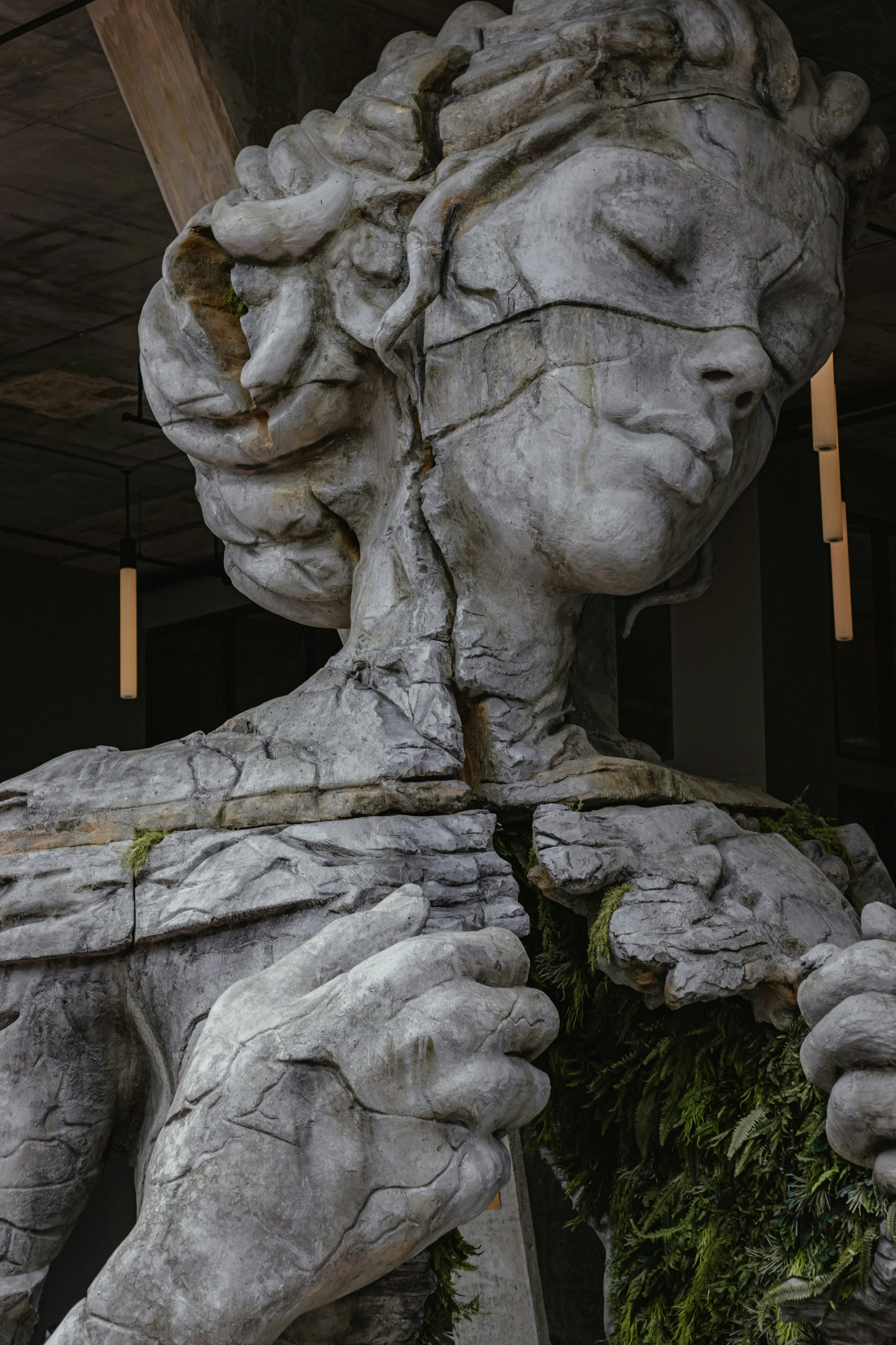 A large stone sculpture of a woman with closed eyes, holding a branch near her neck, with moss and greenery at the base.
