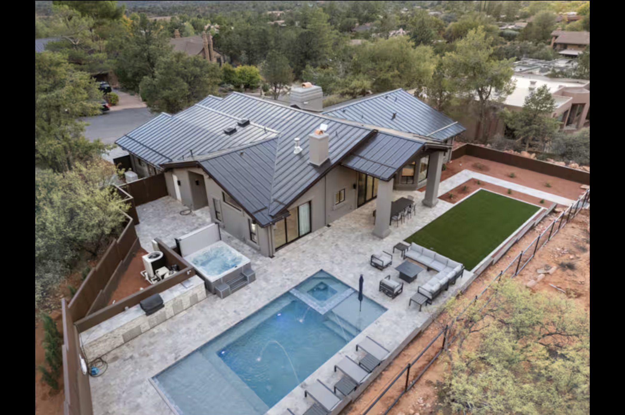Aerial view of a modern house's backyard showing a swimming pool with a jacuzzi, outdoor seating area, artificial grass, and a patio, surrounded by trees and neighboring houses.