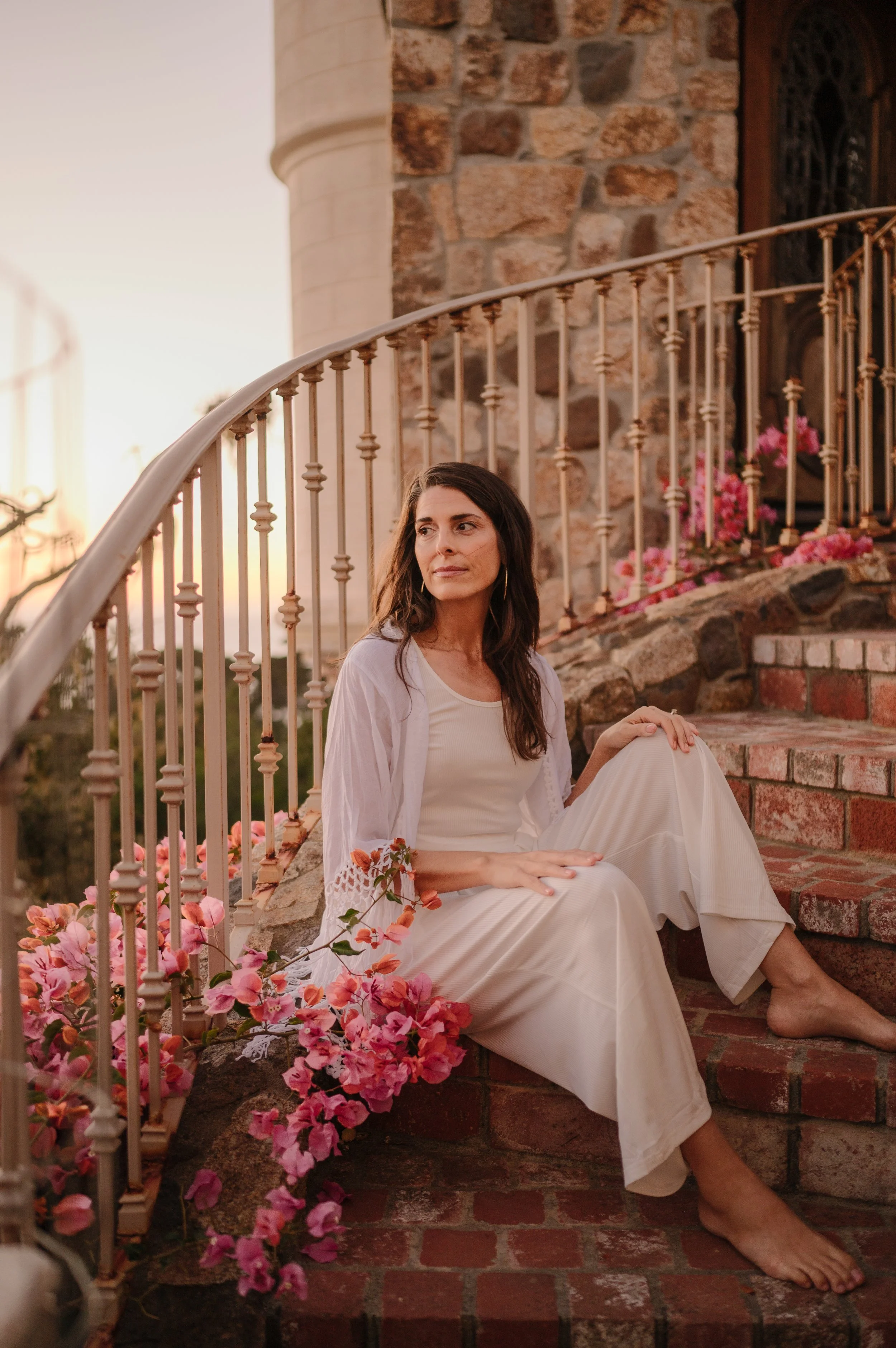 A woman in white attire sitting barefoot on brick steps outside a stone building, surrounded by pink flowers, during sunset.