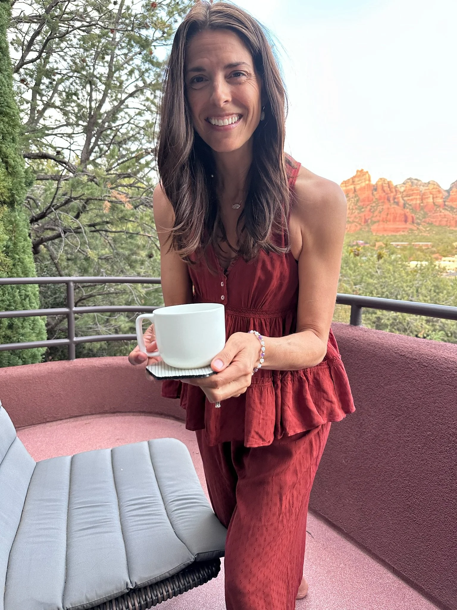 A woman with long brown hair smiling and holding a white coffee mug and saucer on a balcony with trees and red rock formations in the background.