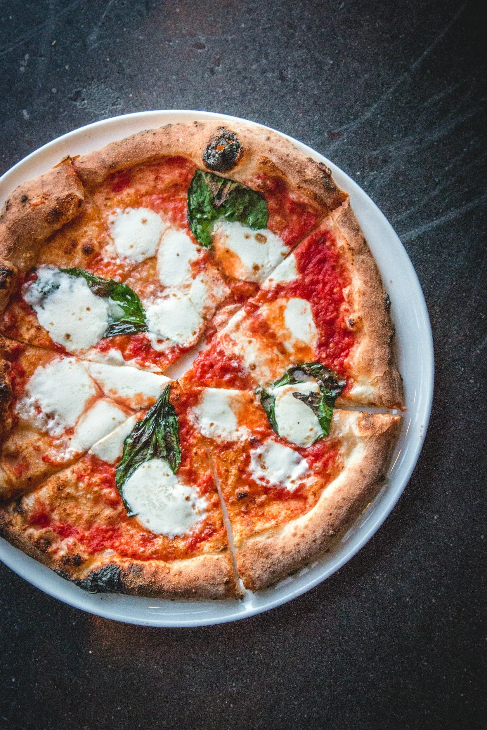 A Margherita pizza with basil leaves, slices of mozzarella, tomato sauce, and a slightly charred crust on a white plate.