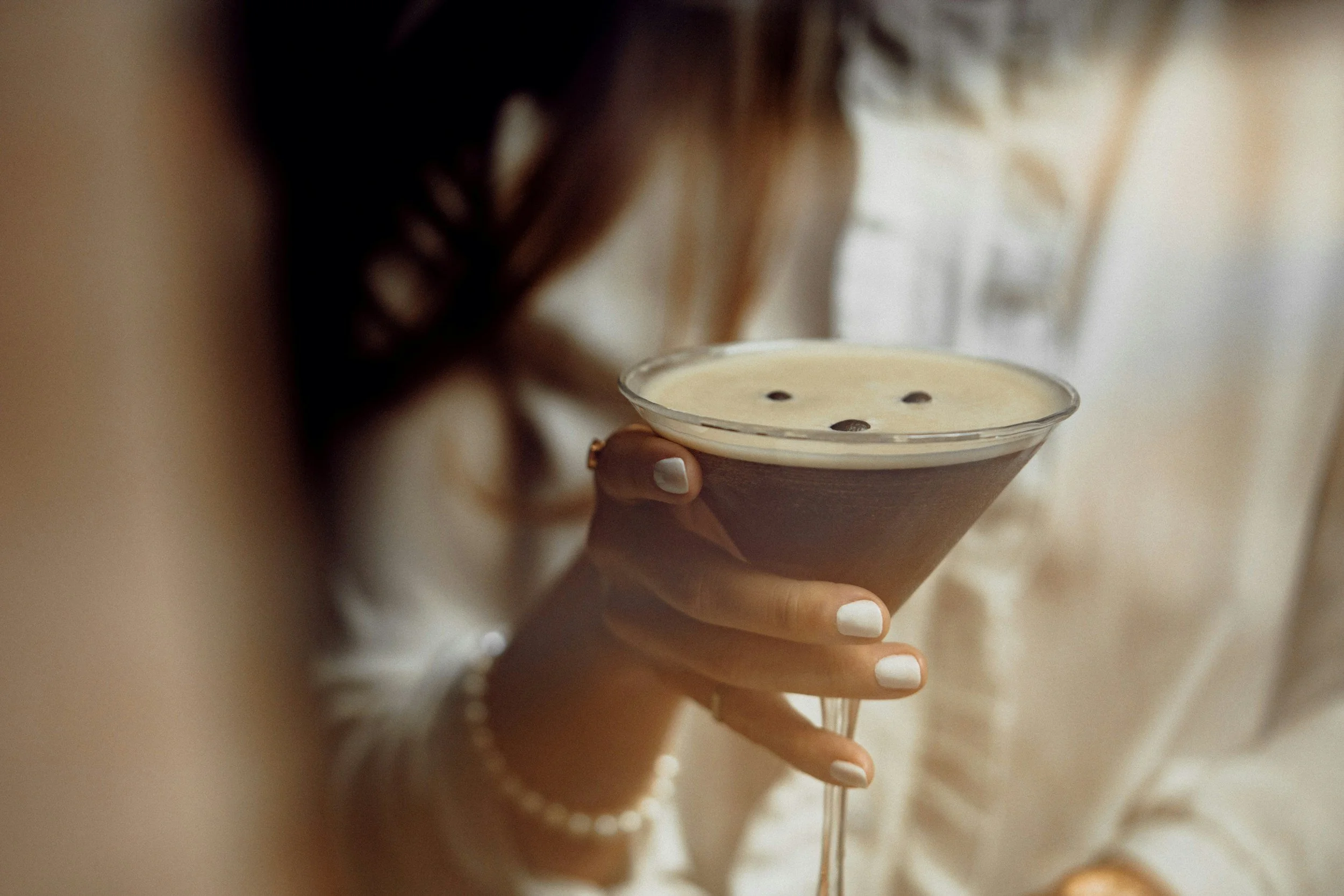 Person holding a dark brown cocktail glass filled with a creamy drink topped with three coffee beans, in a cozy indoor setting.