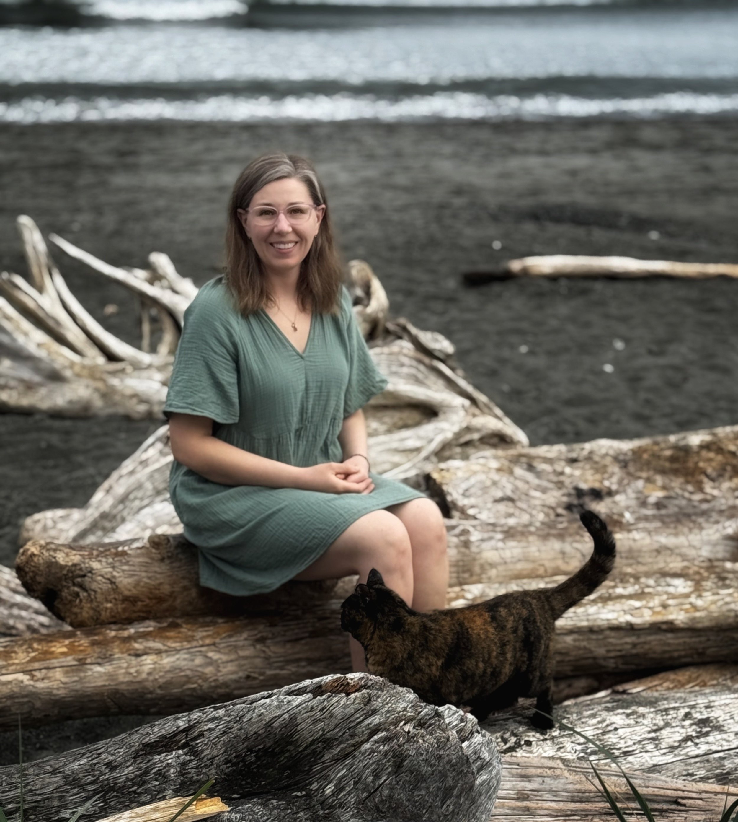 A woman sitting on driftwood on a beach with black sand, looking at the camera, with ocean waves in the background.