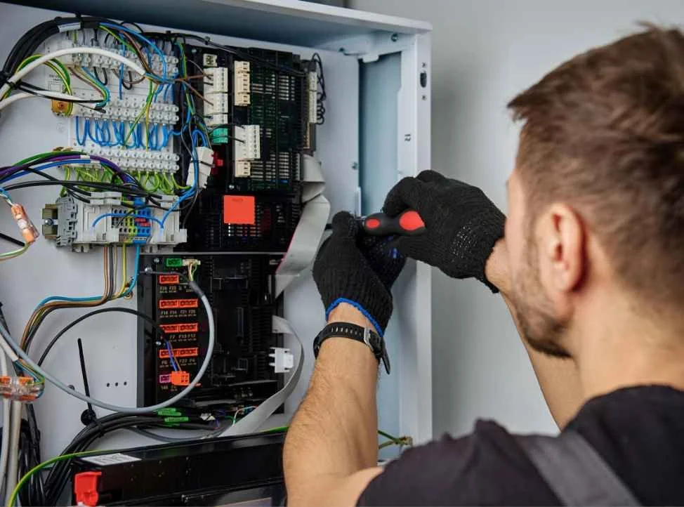 A technician wearing black gloves using a screwdriver to work on an open electrical or control panel with many wires and components inside.