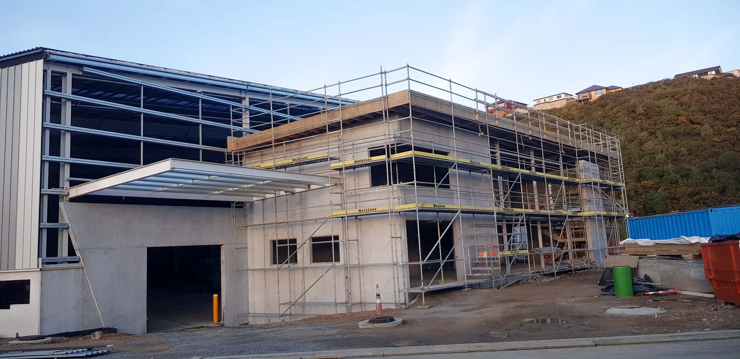 Building under construction with scaffolding, concrete walls, and a large open garage doorway, set against a hillside with residential houses.