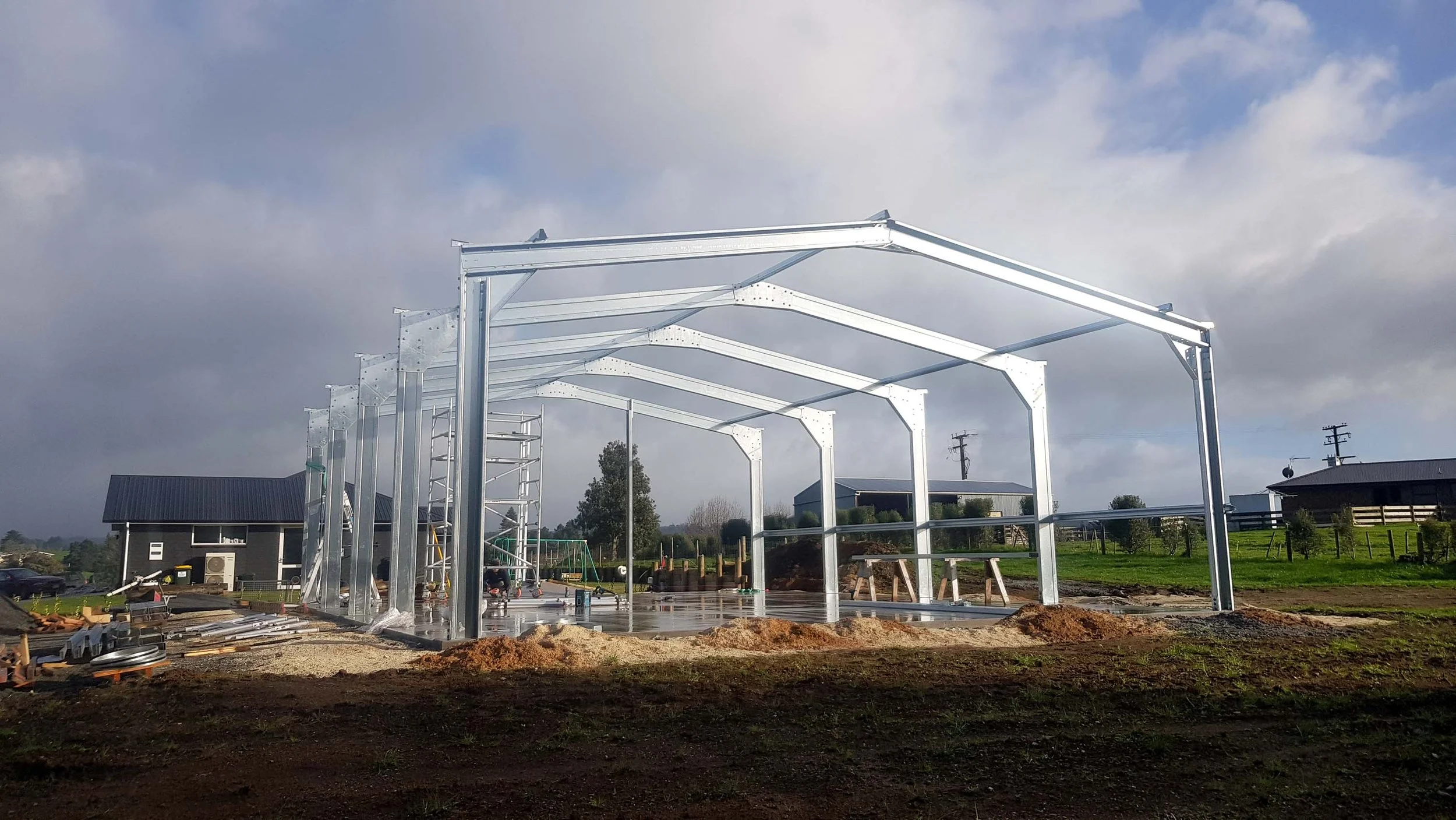 Metal framework being assembled for a building or structure outdoors on dirt ground with construction tools and materials nearby, clear sky with some clouds in the background.