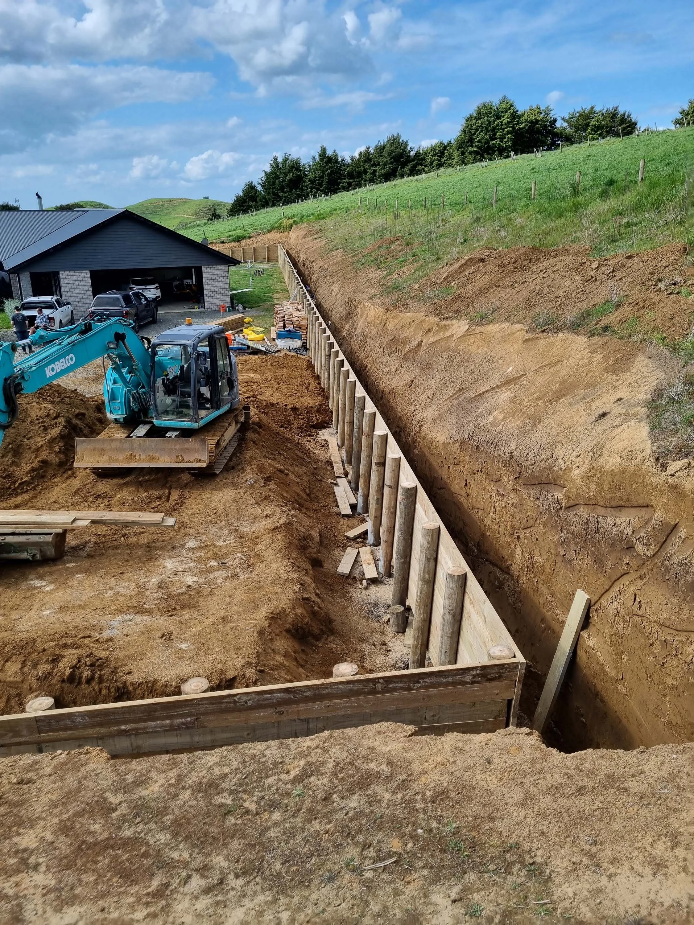 Construction site with a mini excavator, wooden support beams, and a trench along a hillside with green grass and trees.
