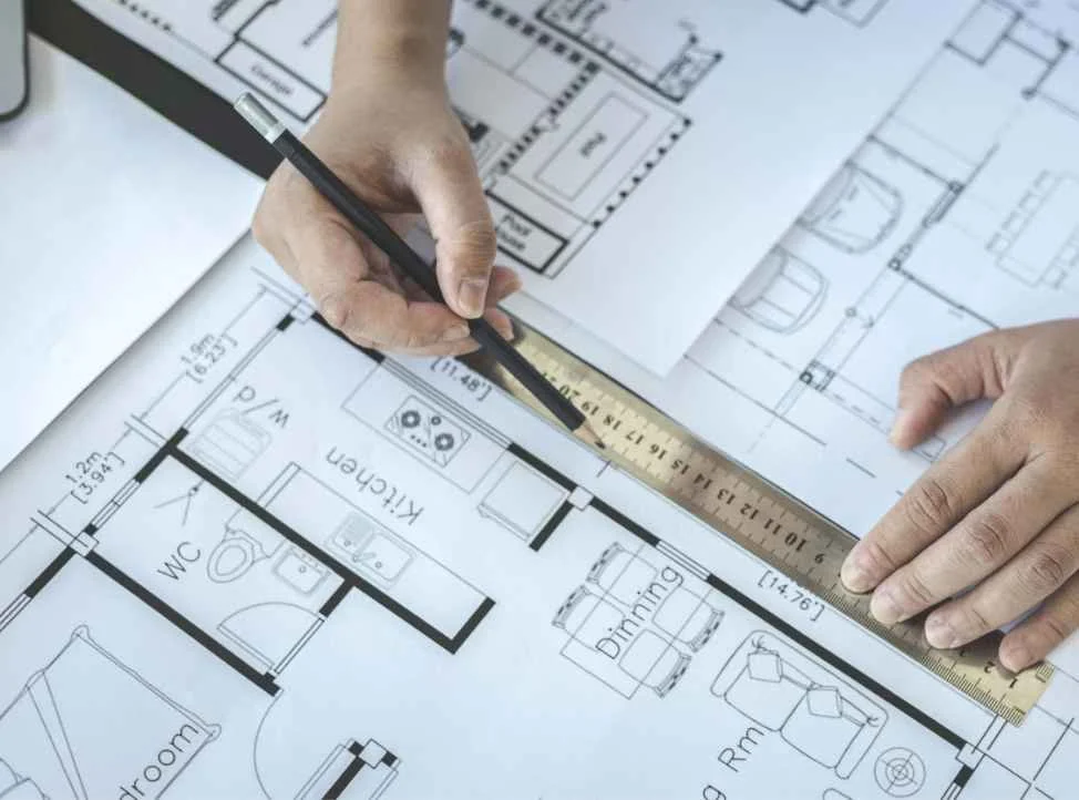 Hands working on an architectural floor plan using a ruler and pencil, with rooms labeled such as kitchen, WC, and dining.