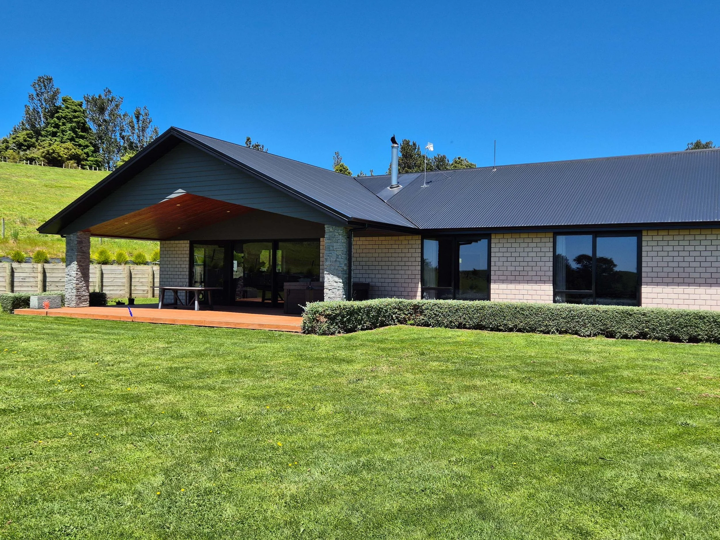 Modern house with stone and brick exterior, large windows, and a wooden deck, surrounded by a neatly maintained lawn and a fenced lush green hillside under a clear blue sky.
