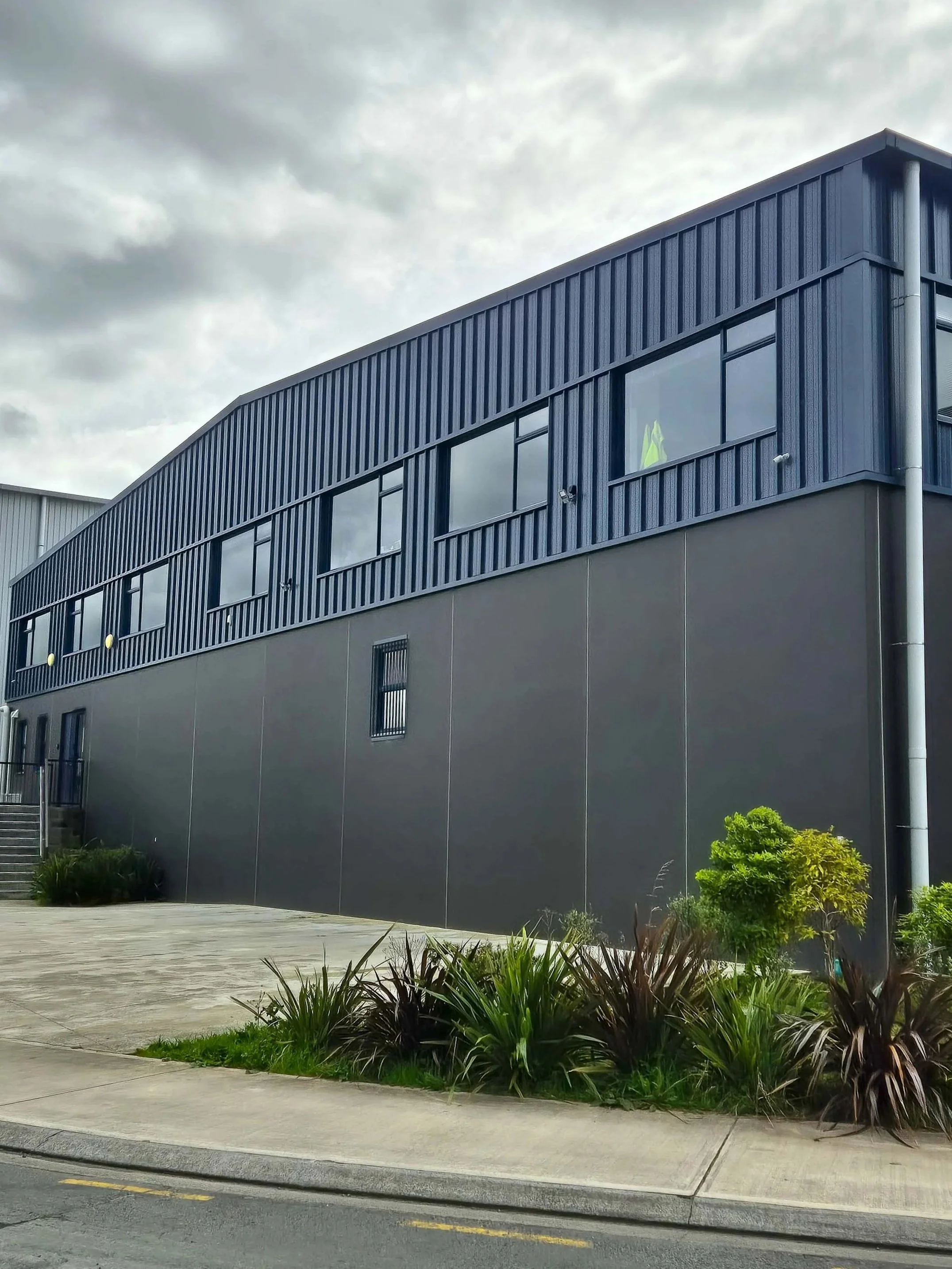 Modern two-story commercial building with blue and gray exterior, several windows, and landscaping with bushes and plants in the foreground.