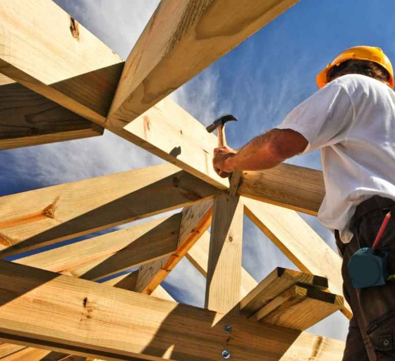 A construction worker wearing a yellow hard hat and white t-shirt hammering a wooden beam into a structure under a blue sky.