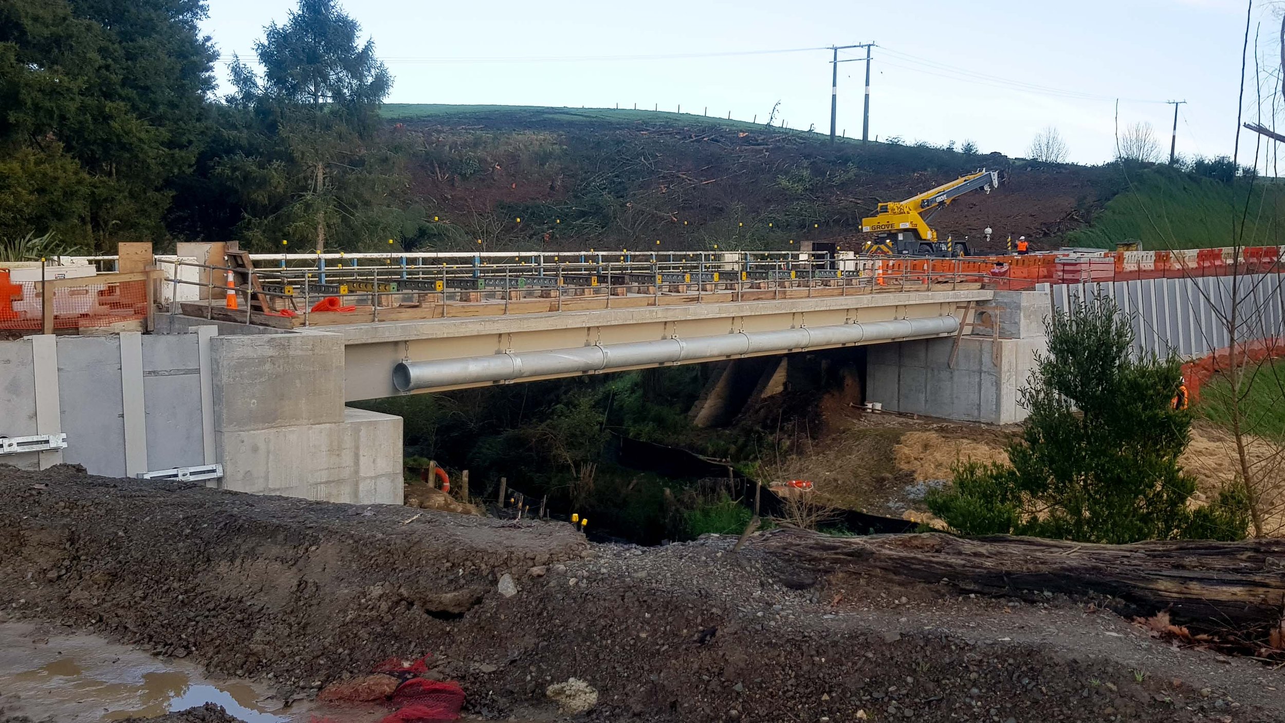 Construction site of a new bridge with visible concrete supports and metal railings, a crane, construction workers, and orange safety barriers amid a landscape with dirt, trees, and utility poles.