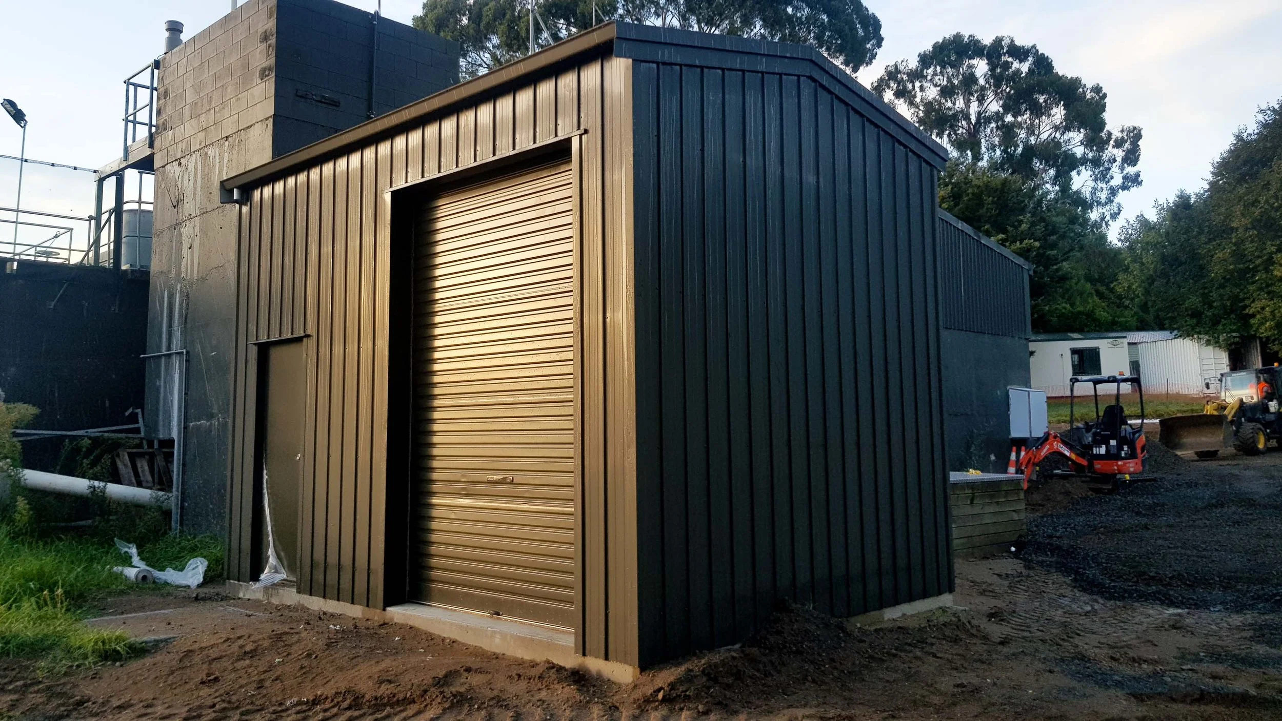 Small black metal shed with beige roll-up door in a construction site with construction equipment and dirt in the background.