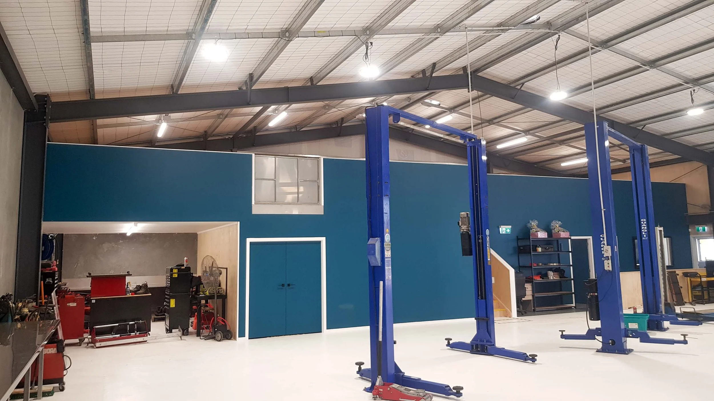 Industrial workshop with blue vehicle lifts, shelving, tools, and equipment. Blue wall with double doors and a window.