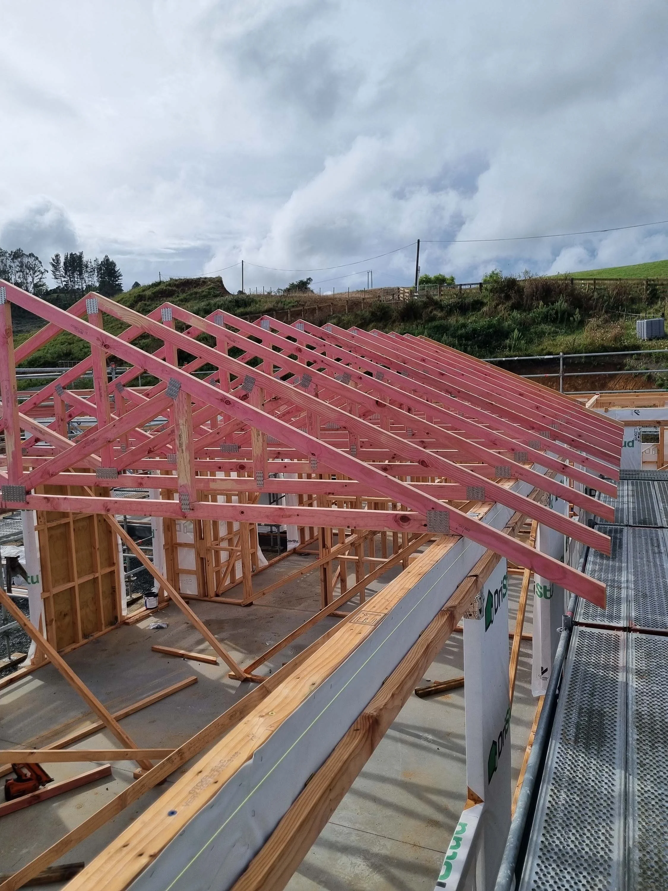 Construction site of a house with pink wooden roof trusses installed, showing framing in progress against a cloudy sky and green hills in the background.