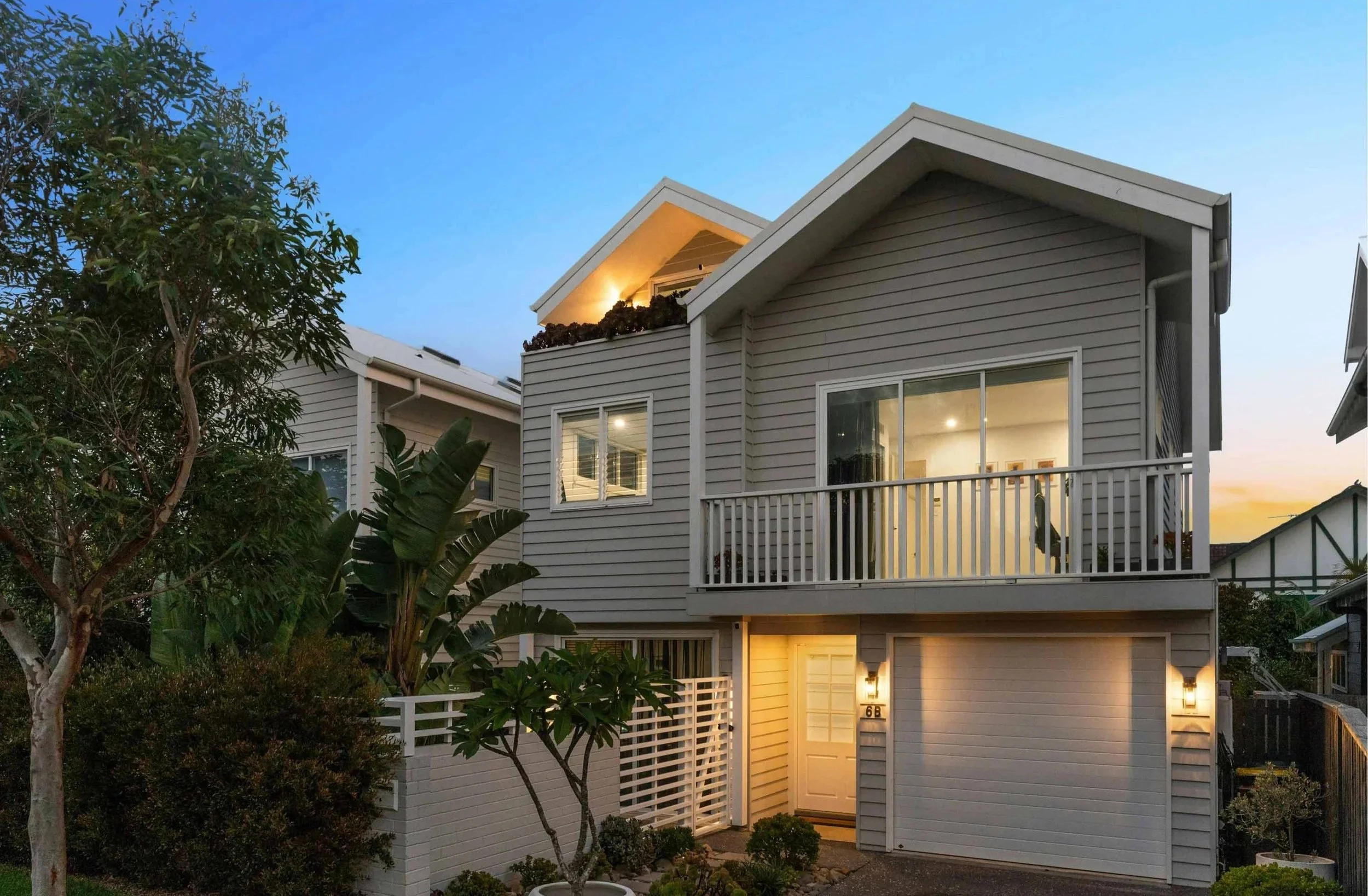 A modern two-story house at dusk with exterior lights on, gray siding, a balcony with glass doors, and lush greenery in the front yard.