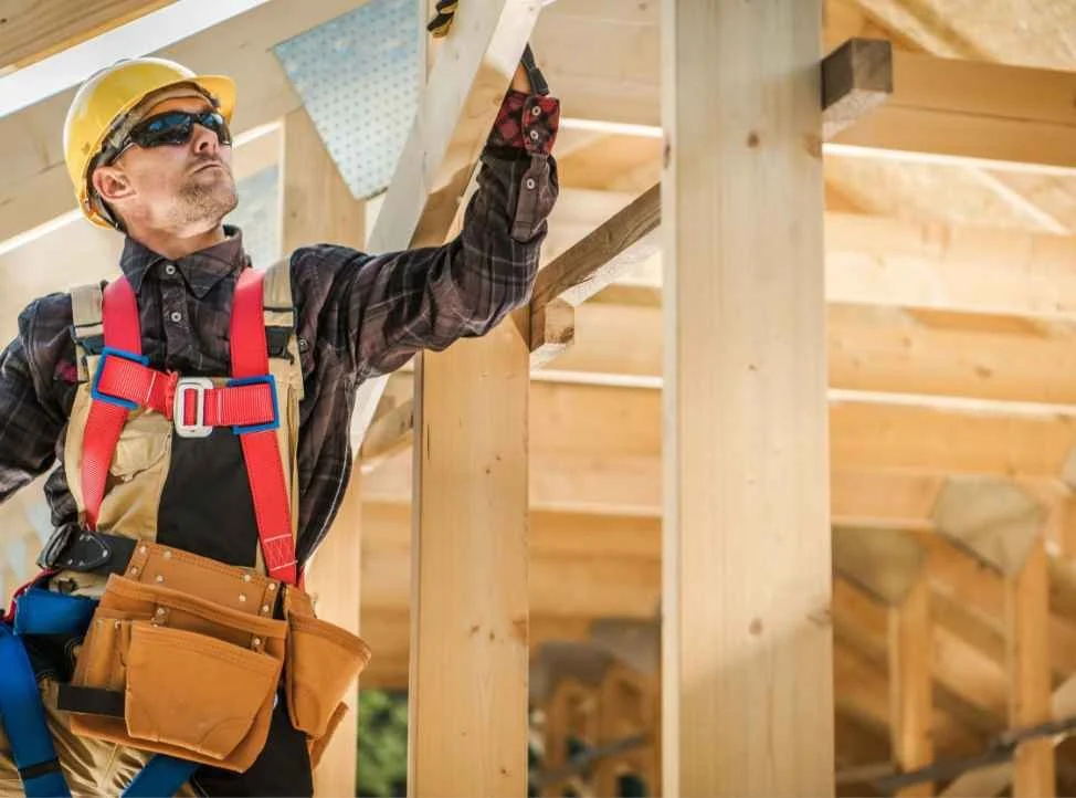 A construction worker wearing a yellow helmet, sunglasses, and a plaid shirt inspecting a wooden structure at a construction site.