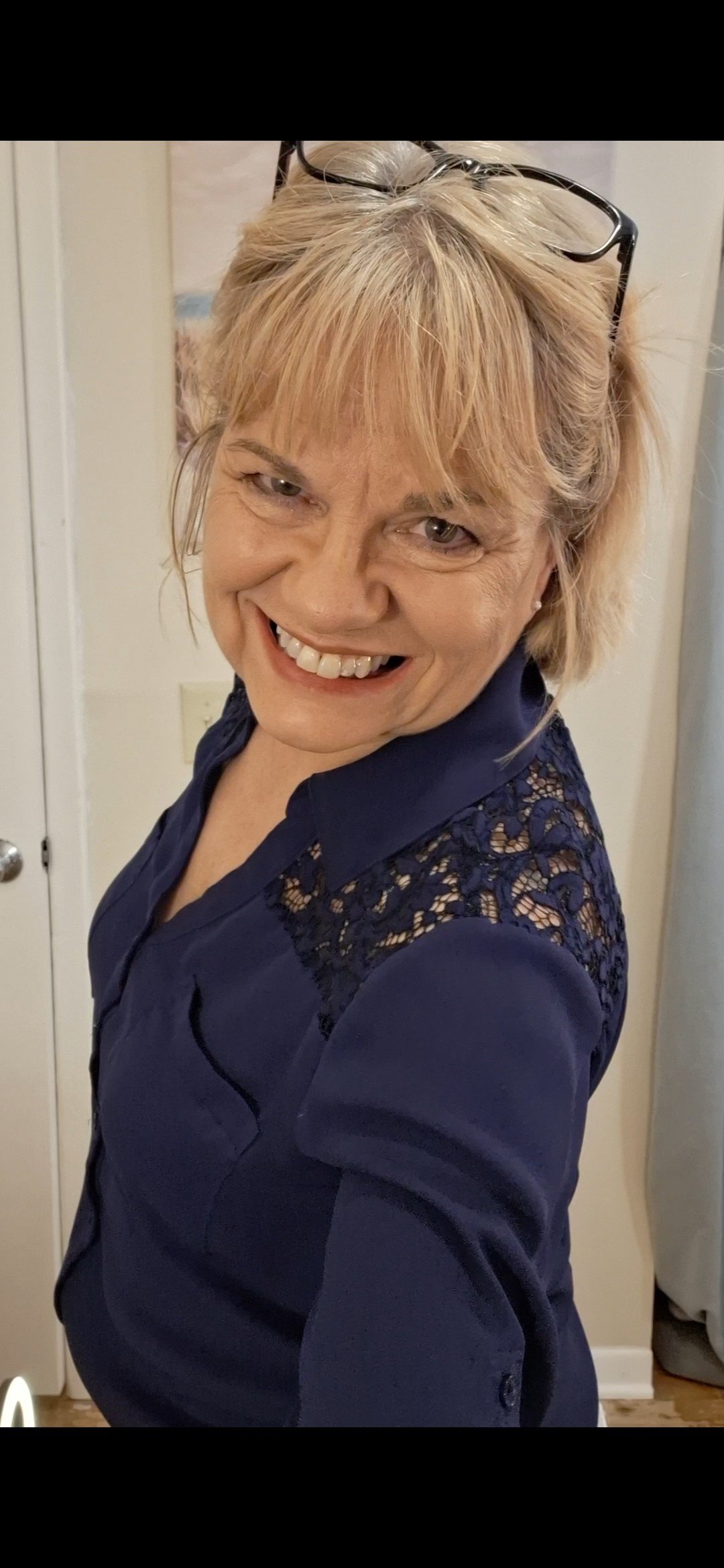A woman with blonde hair, smiling and wearing glasses on her head, dressed in a navy blue top with lace detailing on the shoulders, standing indoors.