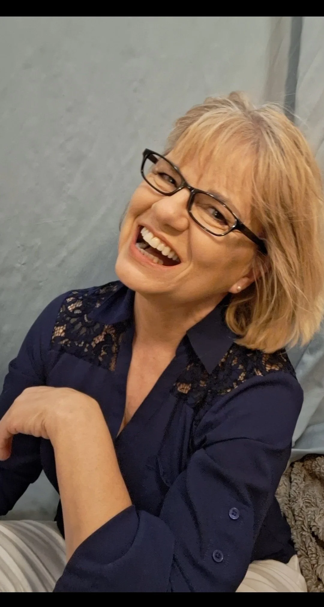 A woman with short blonde hair and glasses smiling broadly, sitting with her chin resting on her hand, wearing a navy blue top with lace details on the shoulders.
