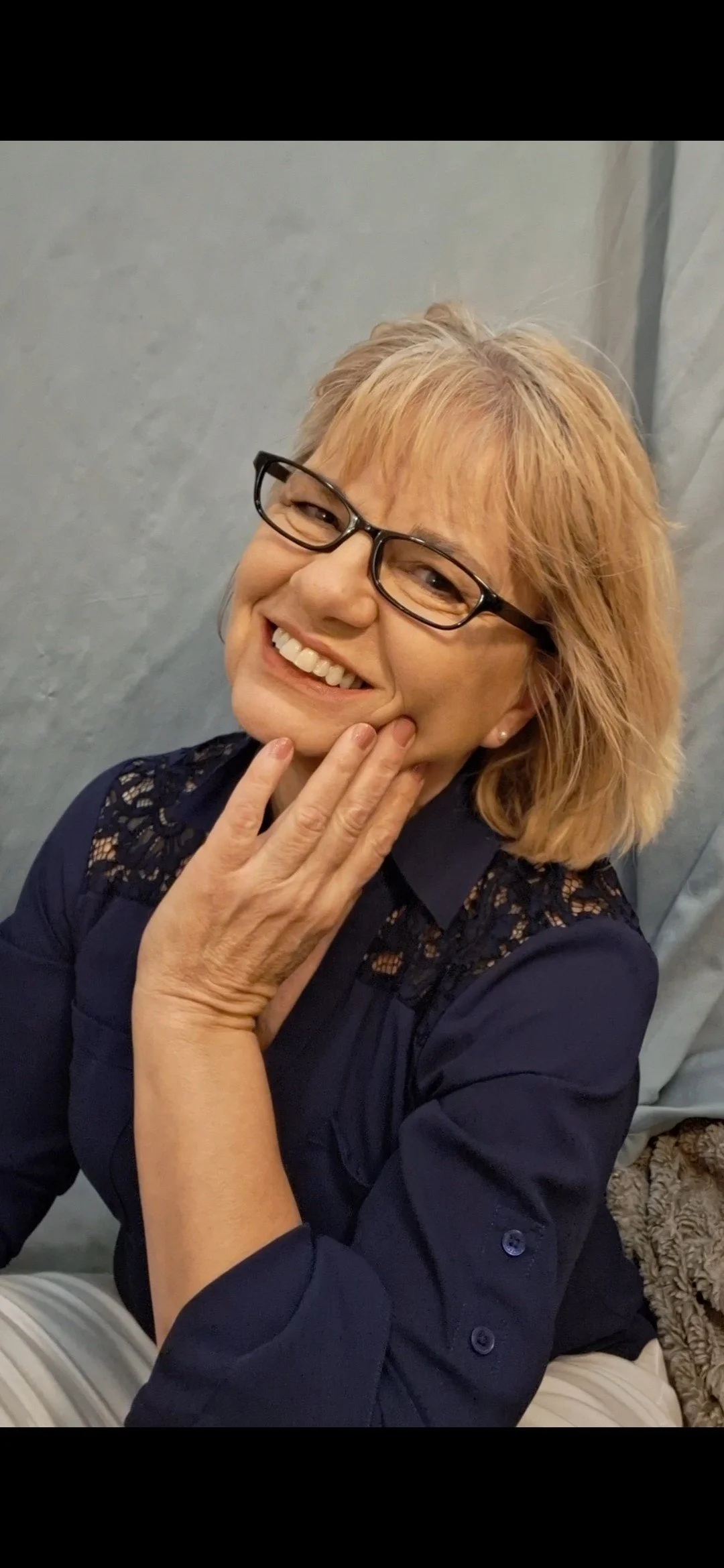 A smiling woman with glasses, blondish hair, and a navy blue top with lace details, posing with her hand on her face against a gray background.