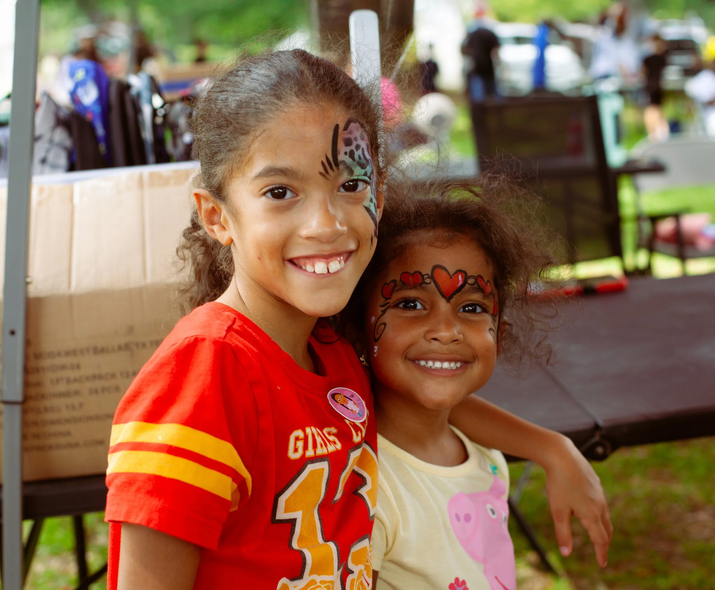 Two young girls with face paint, smiling at an outdoor event. One wears a red shirt with yellow stripes, and the other a yellow shirt with a pink pig design. They have face paintings with hearts and other colorful designs.