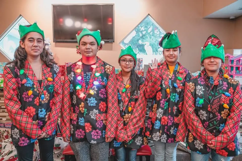 Five children wearing Christmas pajamas and elf hats, with colorful lights and decorations in the background.