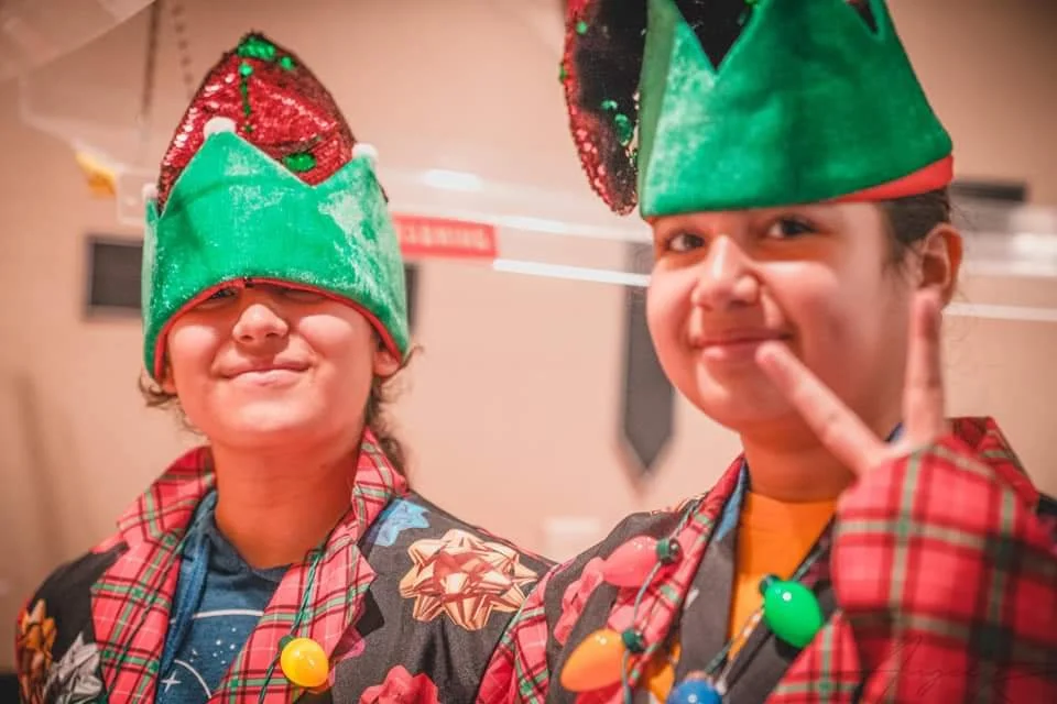 Two children wearing Christmas elf hats and plaid costumes, smiling and making a peace sign with their hands.