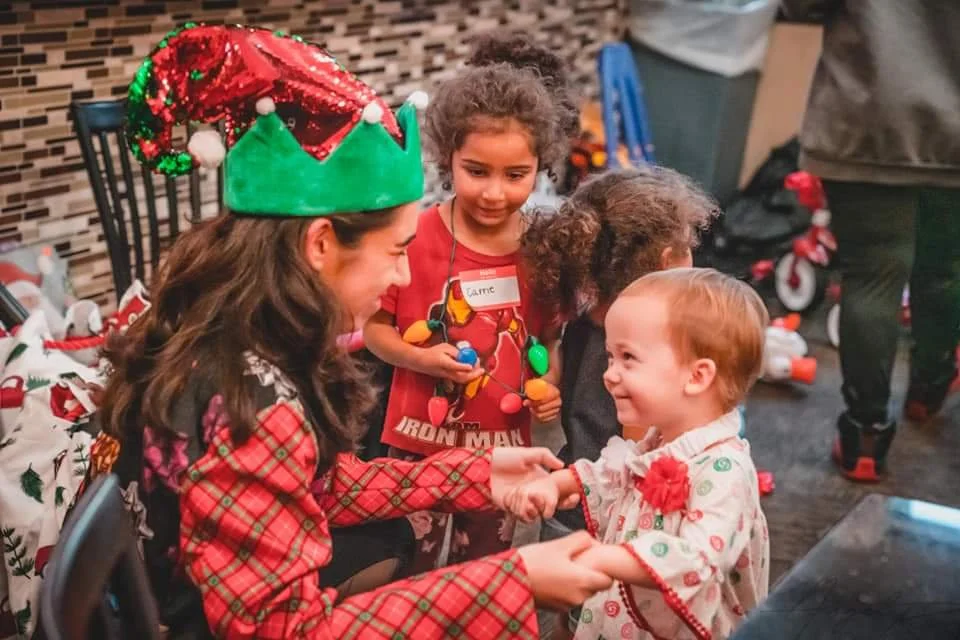 A woman dressed in a Christmas-themed outfit and festive elf hat is smiling and holding a young child's hands. The child is wearing a white pajama top with green and red patterns and a red flower decoration. Behind them, two girls are watching, one w