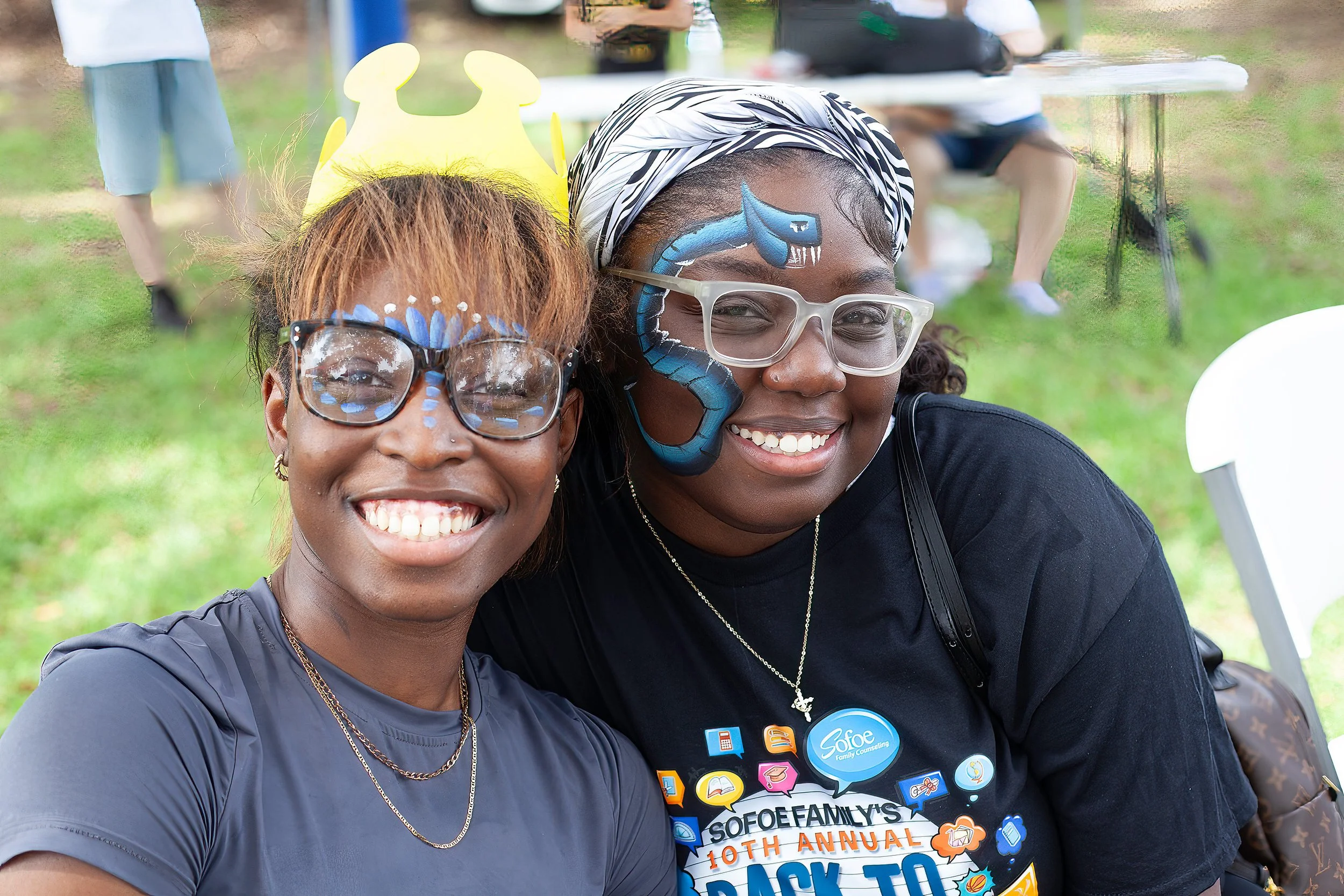 Two women smiling at an outdoor event, with face paint and accessories, one wearing a yellow crown and the other a headscarf, at Sofee Family's 10th annual back to school event.