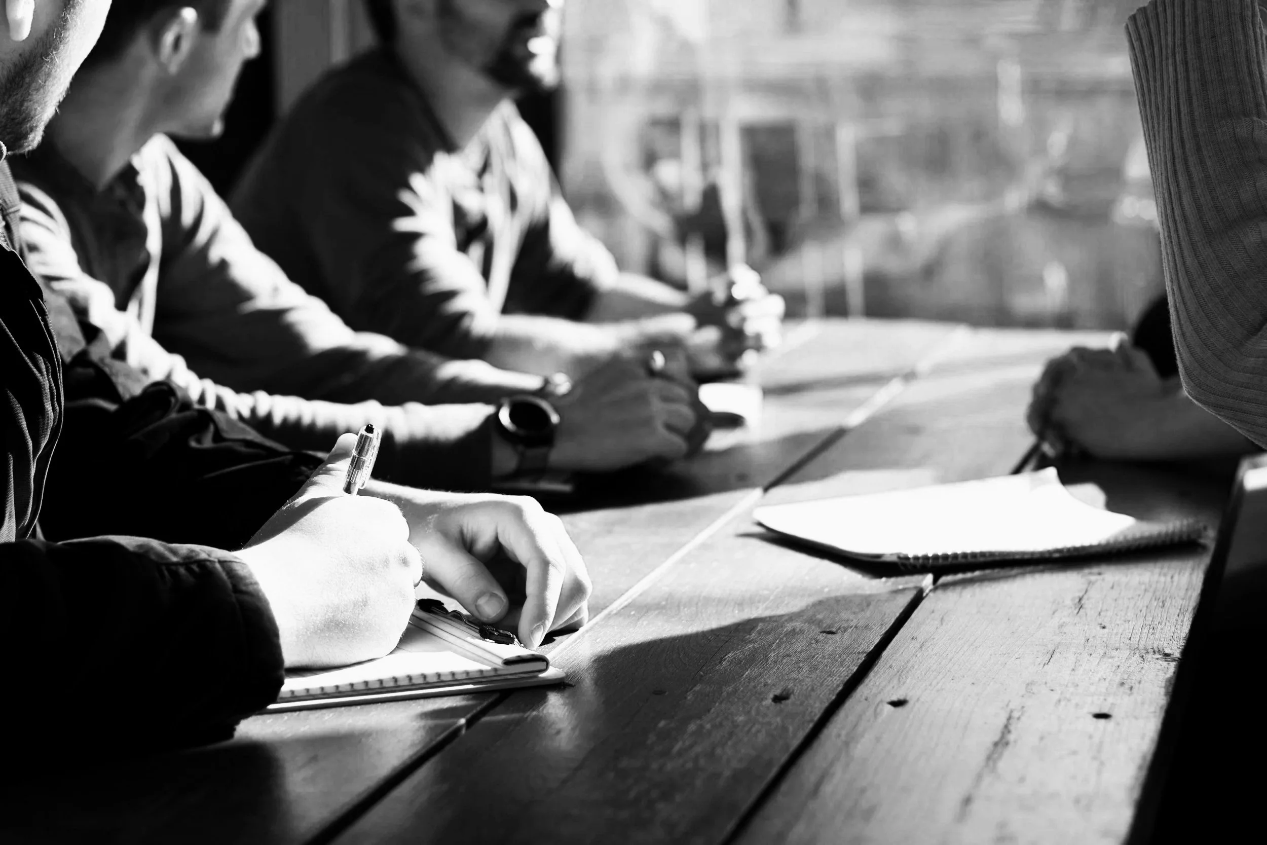 People sitting at a wooden table, taking notes, with notebooks and pens in a meeting or discussion, in black and white.