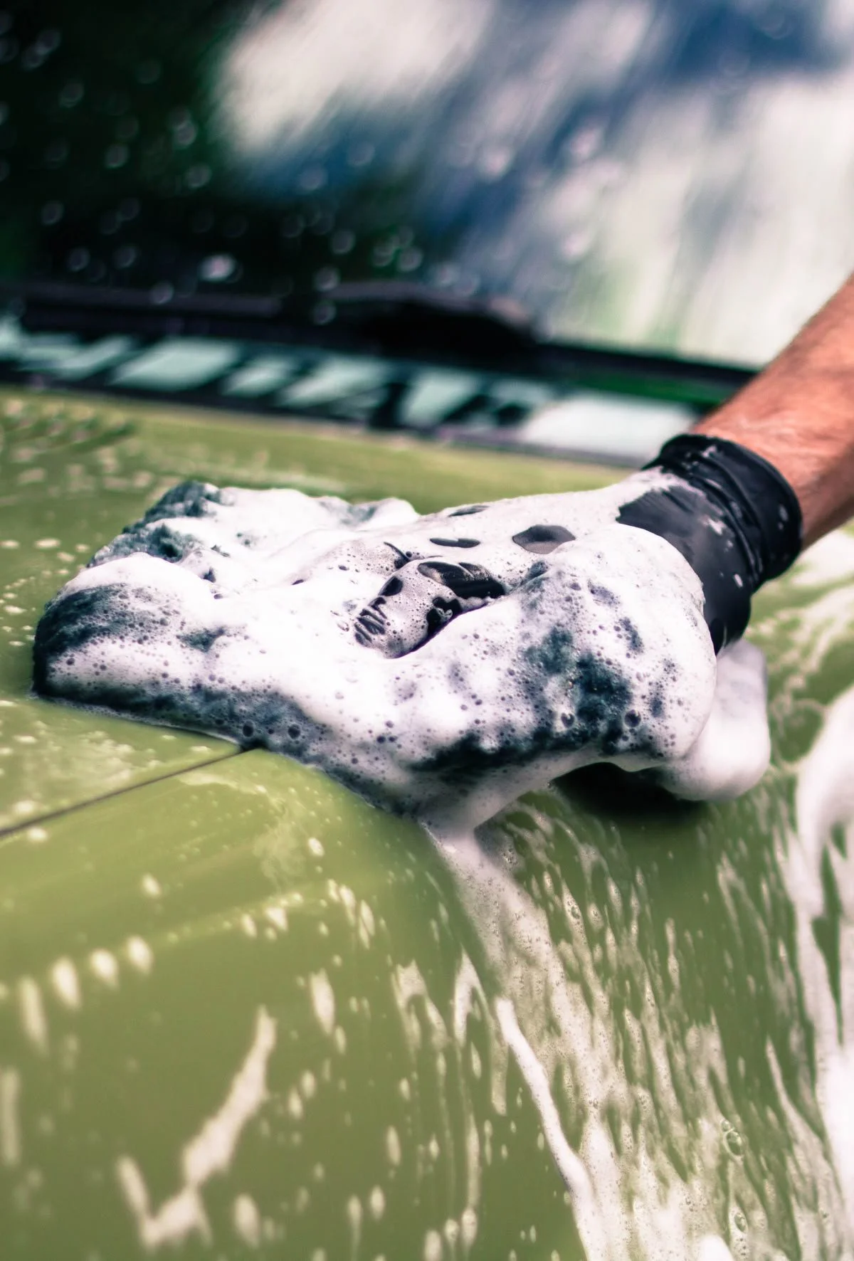 Person wearing a black glove washing a green vehicle with soap and water, creating foam and bubbles.