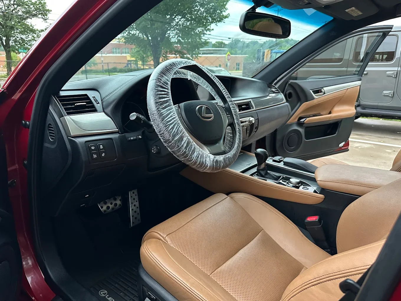 Interior of a Lexus vehicle with a beige leather seat, black dashboard, and steering wheel covered with plastic, parked outside with another vehicle visible through the open door.