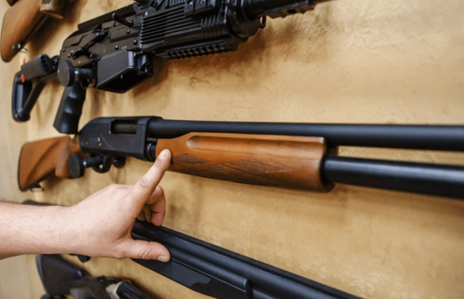 A hand pointing at a rifle with a wooden stock, mounted on a wall with other firearms.