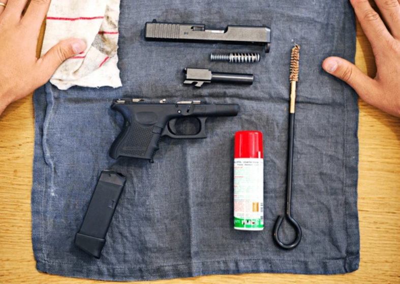 Disassembled handgun with cleaning rod, magazine, cleaning cloth, and can of gun cleaner on a blue cloth