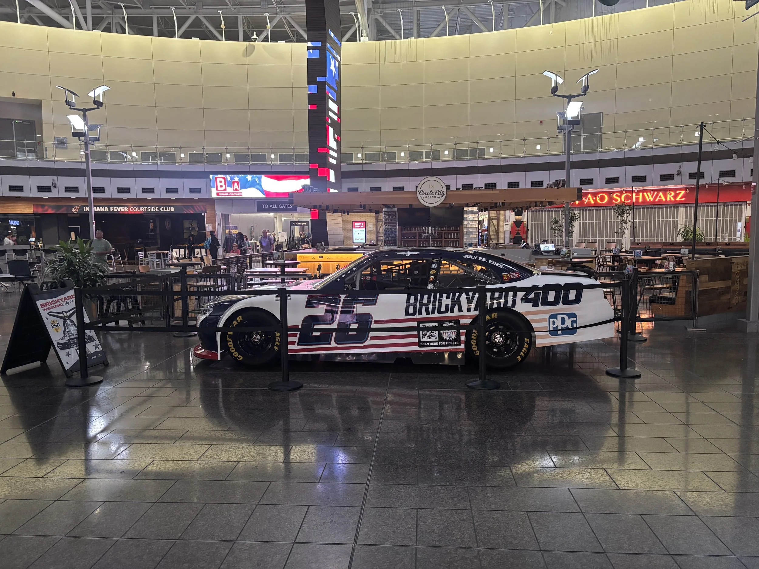 Indoor public space with a race car display in the foreground, people walking, and various signs including one for a beer garden and a club, with high ceiling and bright lighting.
