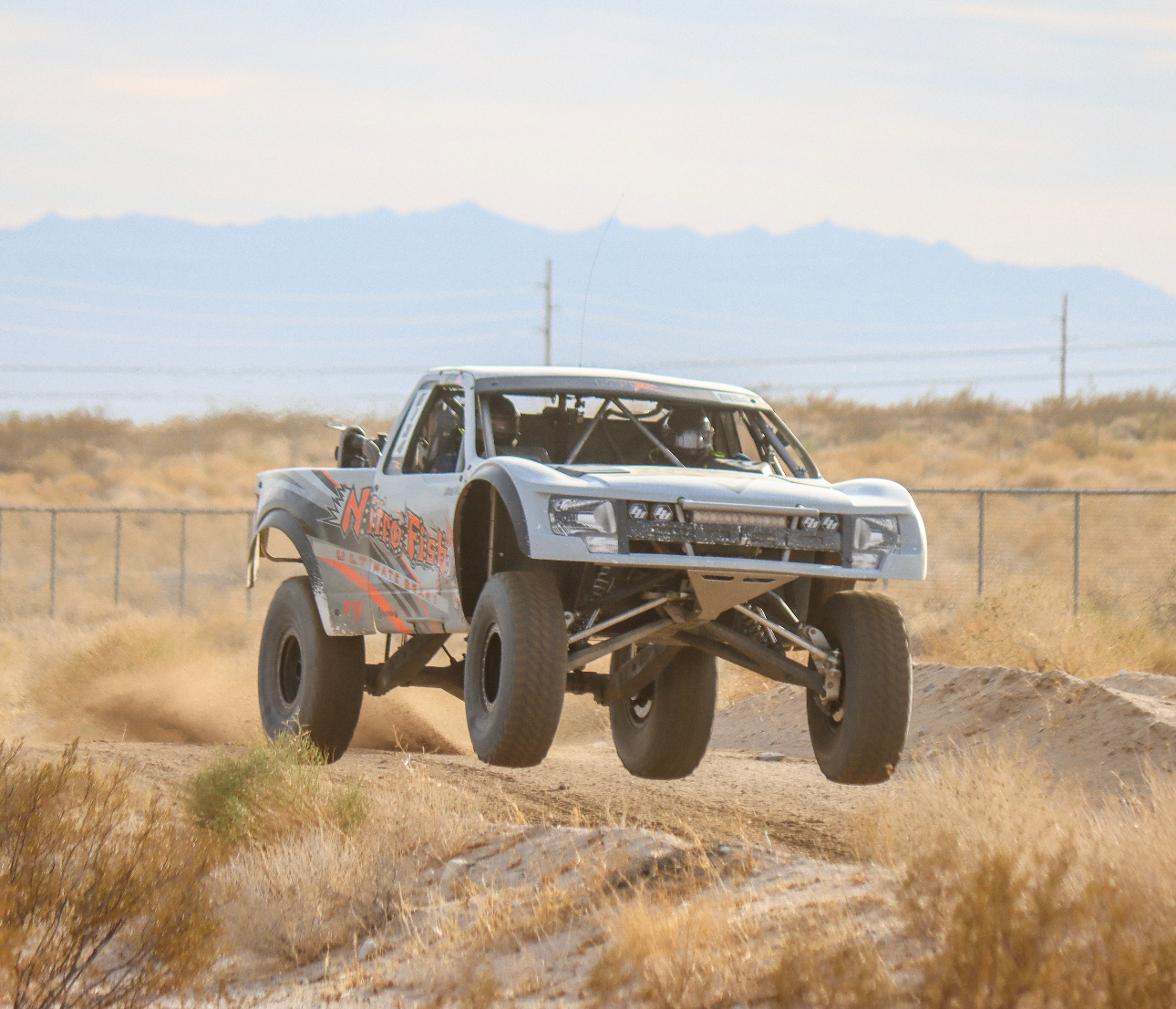A desert off-road race truck labeled Nitro Fish in mid-air jump over dirt and rocks, with mountains and power lines in the background.